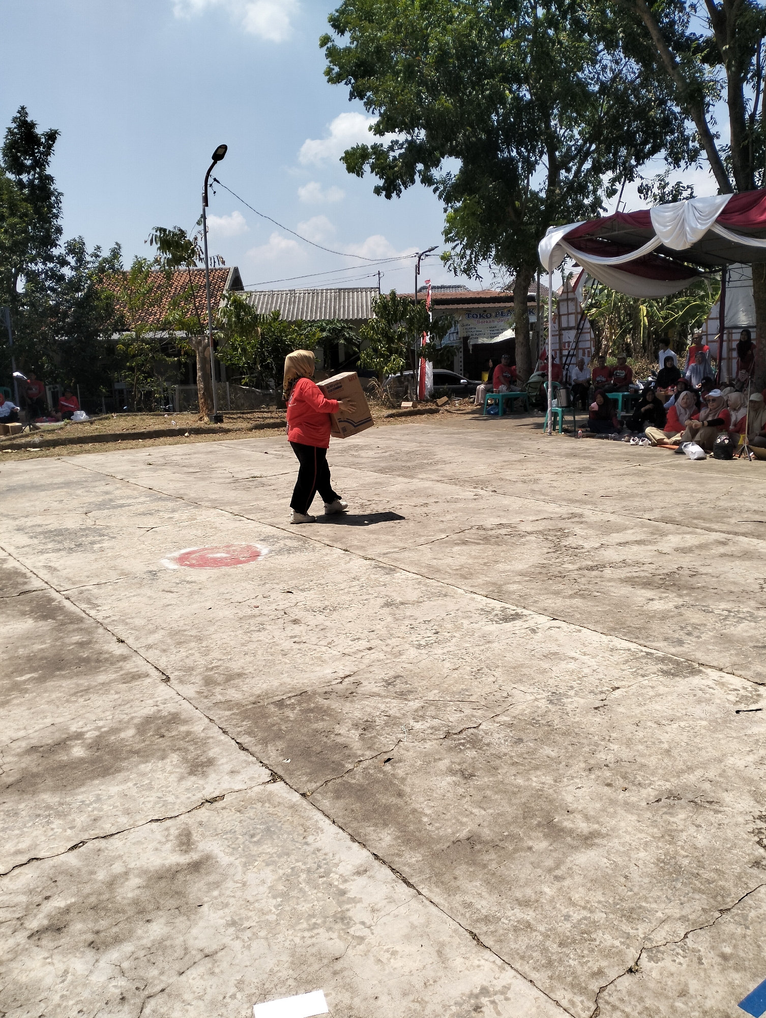 In the city of Semarang, Indonesia, under a clear blue sky with scattered clouds, a young woman wearing a bright orange long-sleeved shirt and dark pants, with a light brown hijab, carries a large cardboard box across a cracked concrete ground. She walks towards the right side of the frame. Behind her, to the left, a streetlamp with a dark fixture stands tall. In the background, a cluster of houses with reddish-brown tile roofs and white-capped roofs are visible, surrounded by lush green trees. To the right, a white and red tent is set up, providing shade. Beneath the tent and in front of it, a group of people are seated on the ground, some on plastic chairs, engaged in an outdoor gathering. A sign with Indonesian text, including "TOKO" (store), is visible on a building in the middle ground. The ground is marked with faint, faded circular shapes, one of which is a red and white target-like design. The overall scene depicts a community event taking place outdoors.