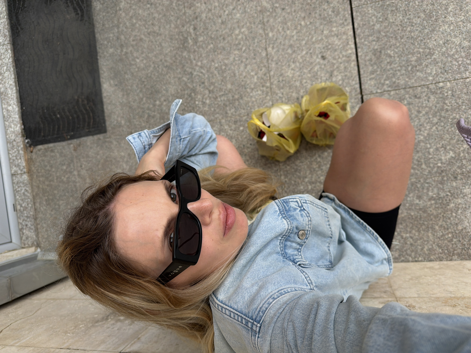 A top-down eye-level photograph shows a woman lying on a tiled floor in what appears to be an outdoor area. She wears a light-wash denim jacket and black knee socks. The woman's sunglasses feature the word "VOGUE" on the temple. To the right and slightly above her are two yellow plastic bags filled with items, possibly food. In the background, there is a dark-colored doormat or similar item and a portion of a window or doorway on the left side of the frame. The tiled floor appears to be a public space, possibly a pedestrian walkway. The location is strongly suggested as Budva, Montenegro, based on the overall appearance and common features of urban areas in that city.