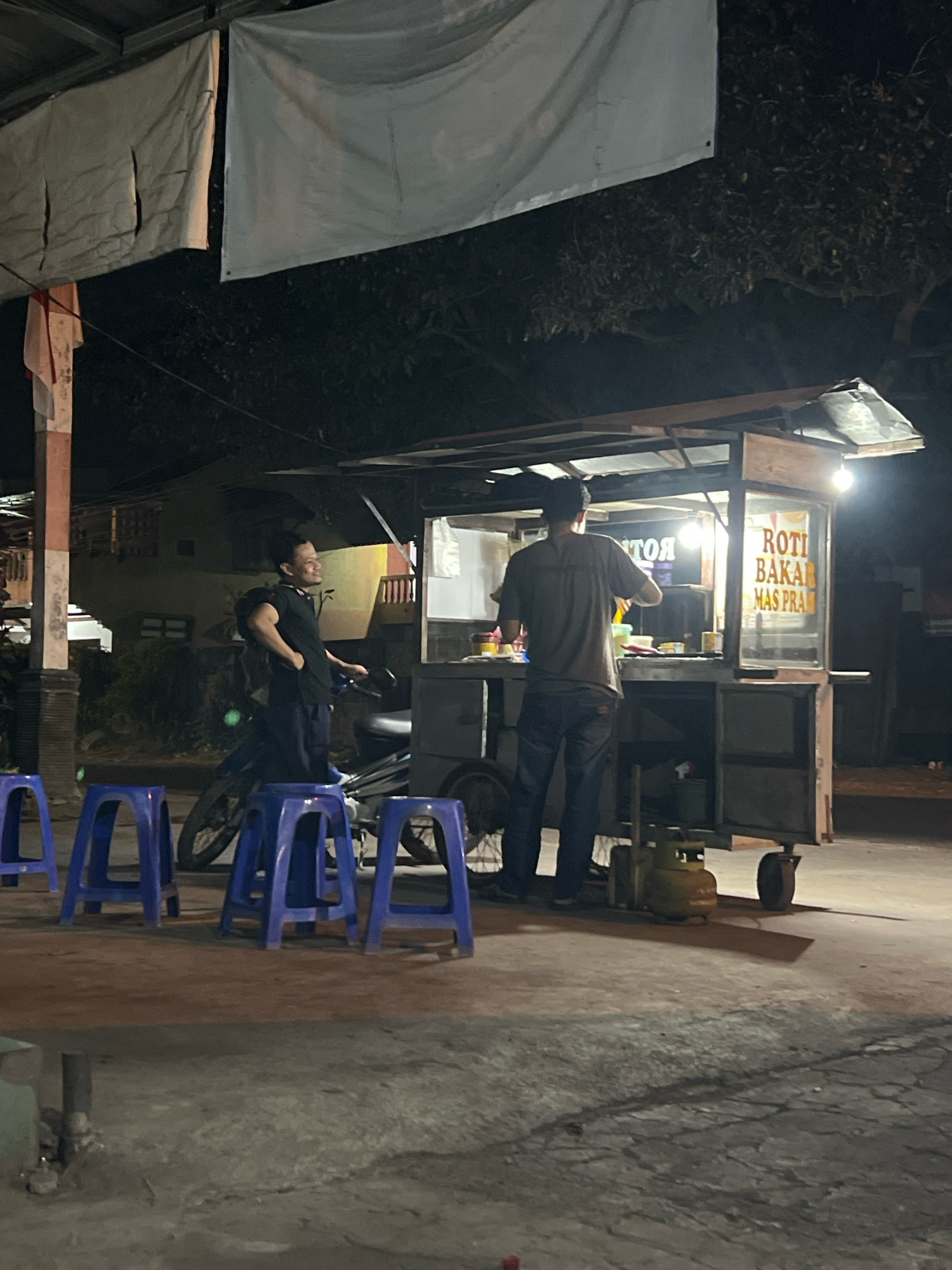 In Tlogoadi, Indonesia, at night, two men are standing at a food cart. The food cart has a sign that reads "ROTI BAKAR MAS PRAS". One man, wearing a black t-shirt and dark pants, is standing near a motorcycle and looking to the right. The other man, wearing a grey t-shirt and dark pants, is facing the cart, working. The cart is illuminated by a bright light. In the foreground, there are several blue plastic stools. The scene appears to be on a street or paved area, with trees and a building visible in the background. A gas cylinder is attached to the cart.