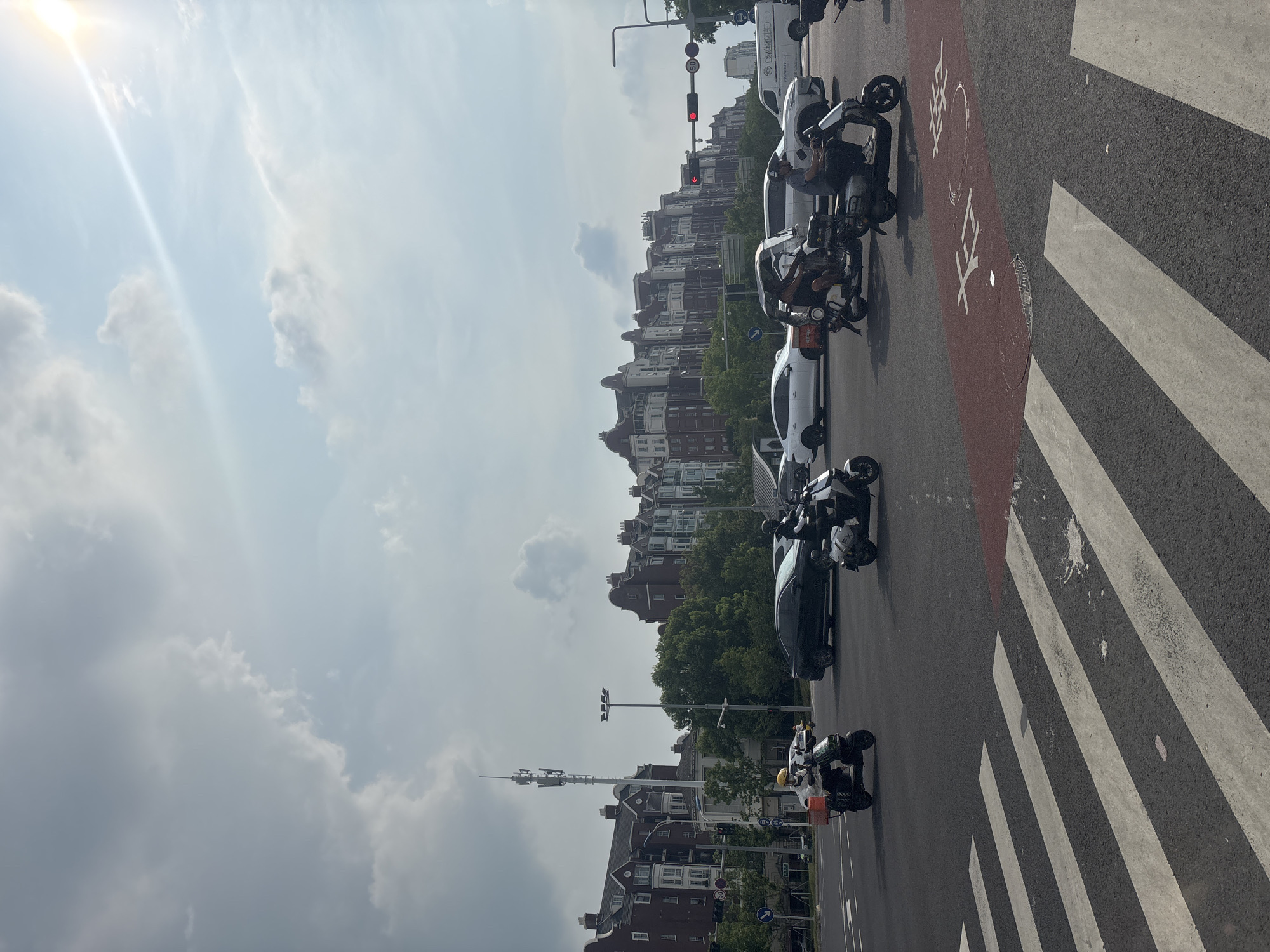 The image depicts a busy intersection in Suzhou Industrial Park, China, on a bright, sunny day with some clouds. Several vehicles, including scooters and cars, are navigating the intersection. Two individuals are riding scooters in the foreground, one wearing a yellow helmet and the other a black helmet. In the background, a row of distinct, multi-story buildings with red brick facades and white accents lines the street. A tall communication tower stands prominently on the left side of the image, and trees are visible between the buildings and the road. Traffic lights are visible, with a red light illuminated. A pedestrian crossing is marked on the asphalt. A white van with Chinese lettering is also present in the scene.