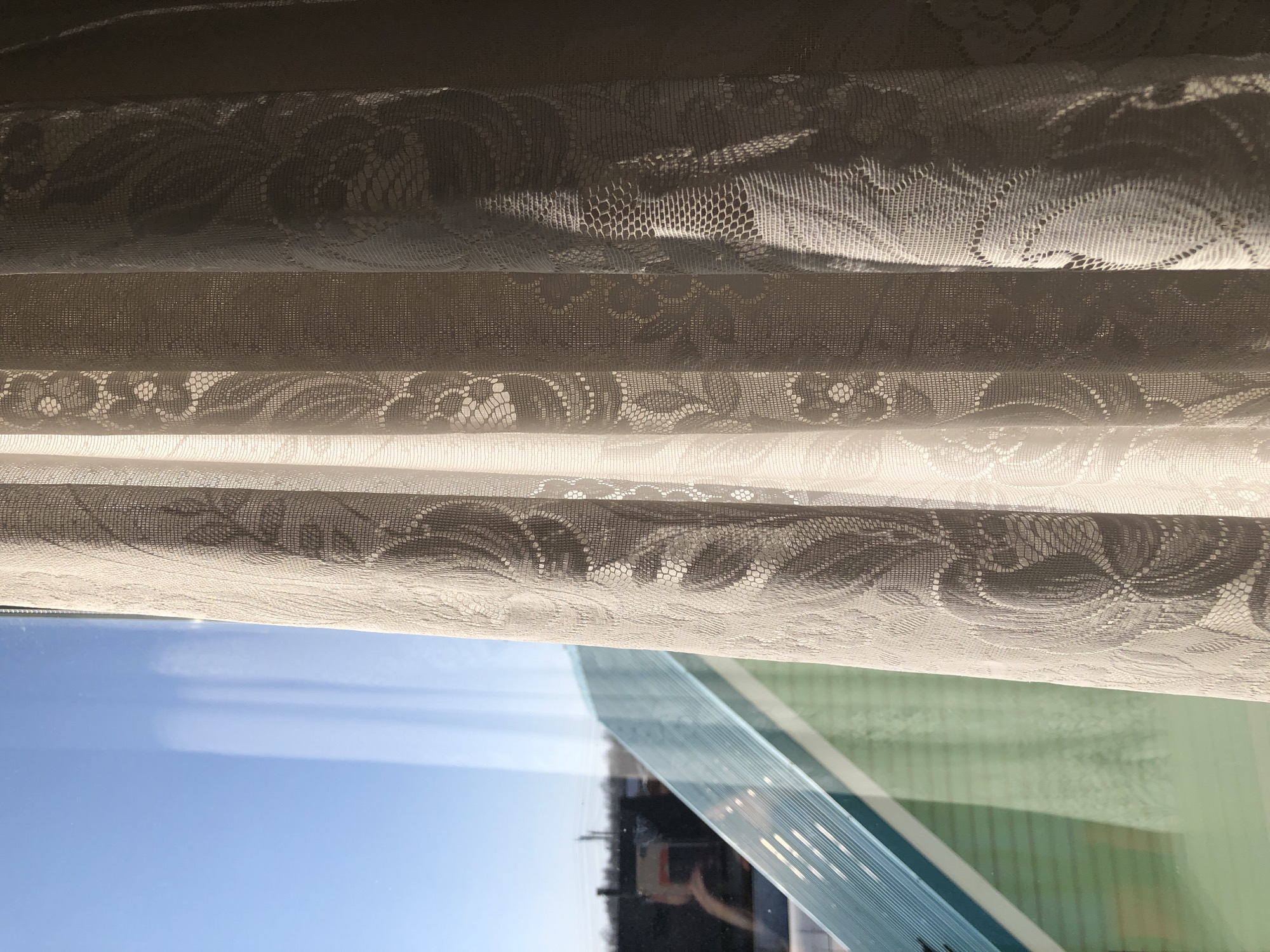 The image depicts a view from inside a room looking out through a window. The foreground is dominated by translucent, patterned lace curtains, folded in layers. Beyond the curtains, the window reflects a partly clear blue sky, a building with what appears to be a green exterior, and the vague silhouette of a person holding a mobile device in the window's reflection. The reflection suggests an urban or suburban area, potentially of Noviy, Russia.