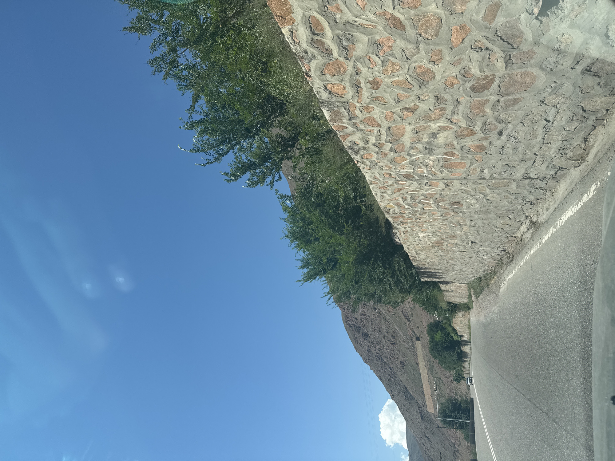 A paved road curves to the left, bordered by a rough stone wall on the right. Beyond the wall, lush green trees grow against a backdrop of steep, rocky brown mountains. A white car is visible driving away from the camera on the road. The sky is a clear, bright blue with a few wispy clouds. The scene suggests a rural or mountainous area, potentially near Cholok, Kyrgyzstan, given the rugged terrain and vegetation.