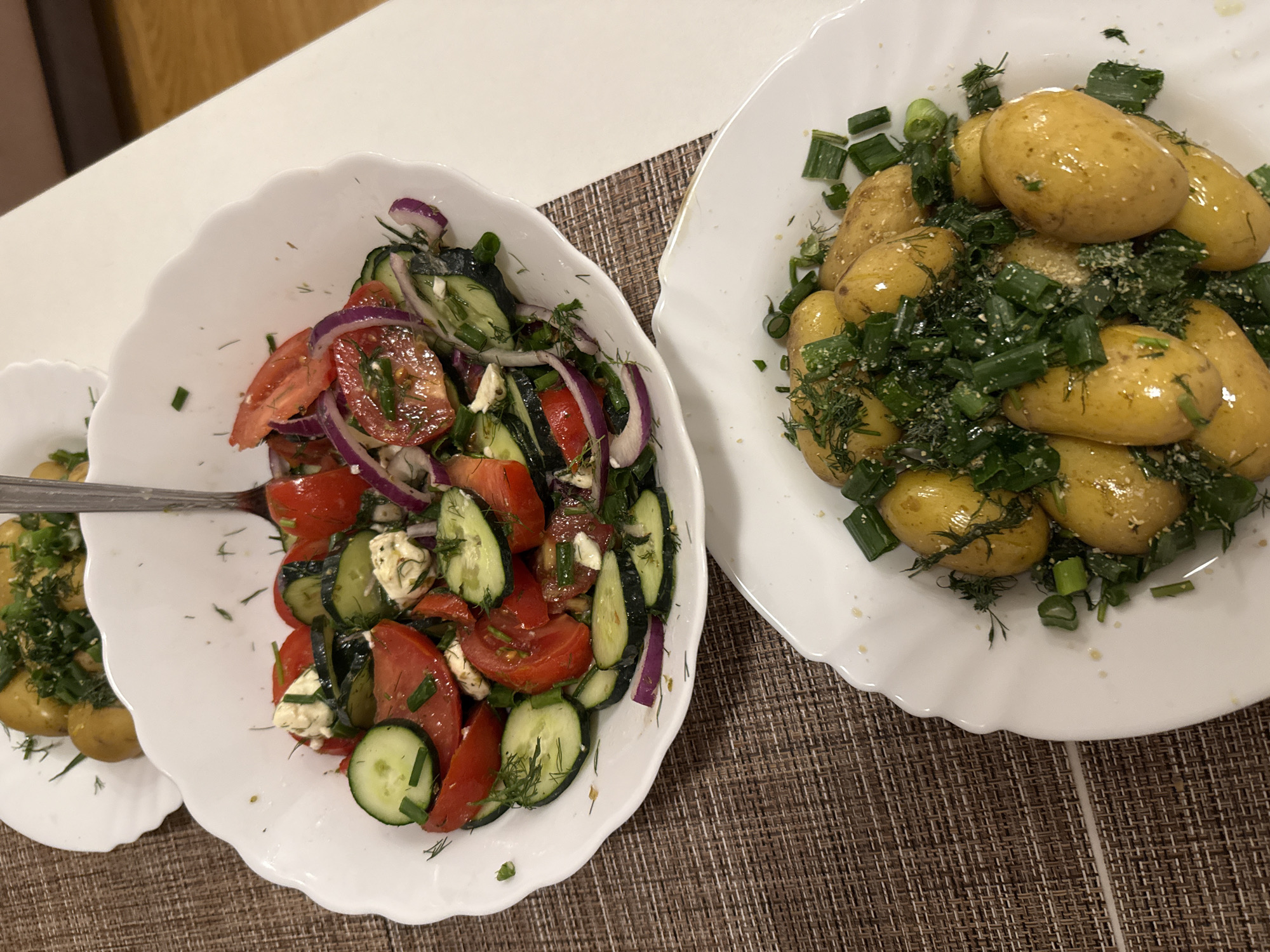 Two white, scalloped-edge plates of food are shown on a brown, woven placemat. The plate in the foreground contains small, whole, golden-yellow potatoes topped with chopped green onions and dill. The plate in the background holds a salad made with sliced tomatoes, cucumbers, red onion, and feta cheese, also garnished with dill and green onions. A fork is resting in the salad. The background is blurred and appears to be an indoor setting.
