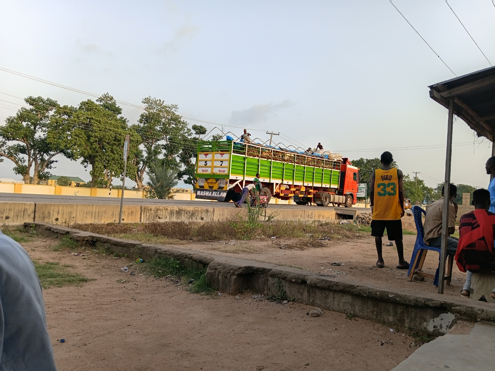 The image depicts a scene in Jalingo, Nigeria, featuring a large green and red truck laden with goods, likely agricultural produce, on a road. Several individuals are visible on top of the truck, seemingly securing the cargo. In the foreground, a group of people are gathered near a rudimentary shelter with a corrugated iron roof. One person stands wearing a yellow tank top with the number "33" and the word "BIRD" on it, while others are seated on chairs and a block. The environment appears to be a roadside area with dusty ground, sparse vegetation, and concrete barriers. Power lines crisscross the sky, indicating the presence of infrastructure. The overall impression is one of everyday life and commerce in a Nigerian setting.