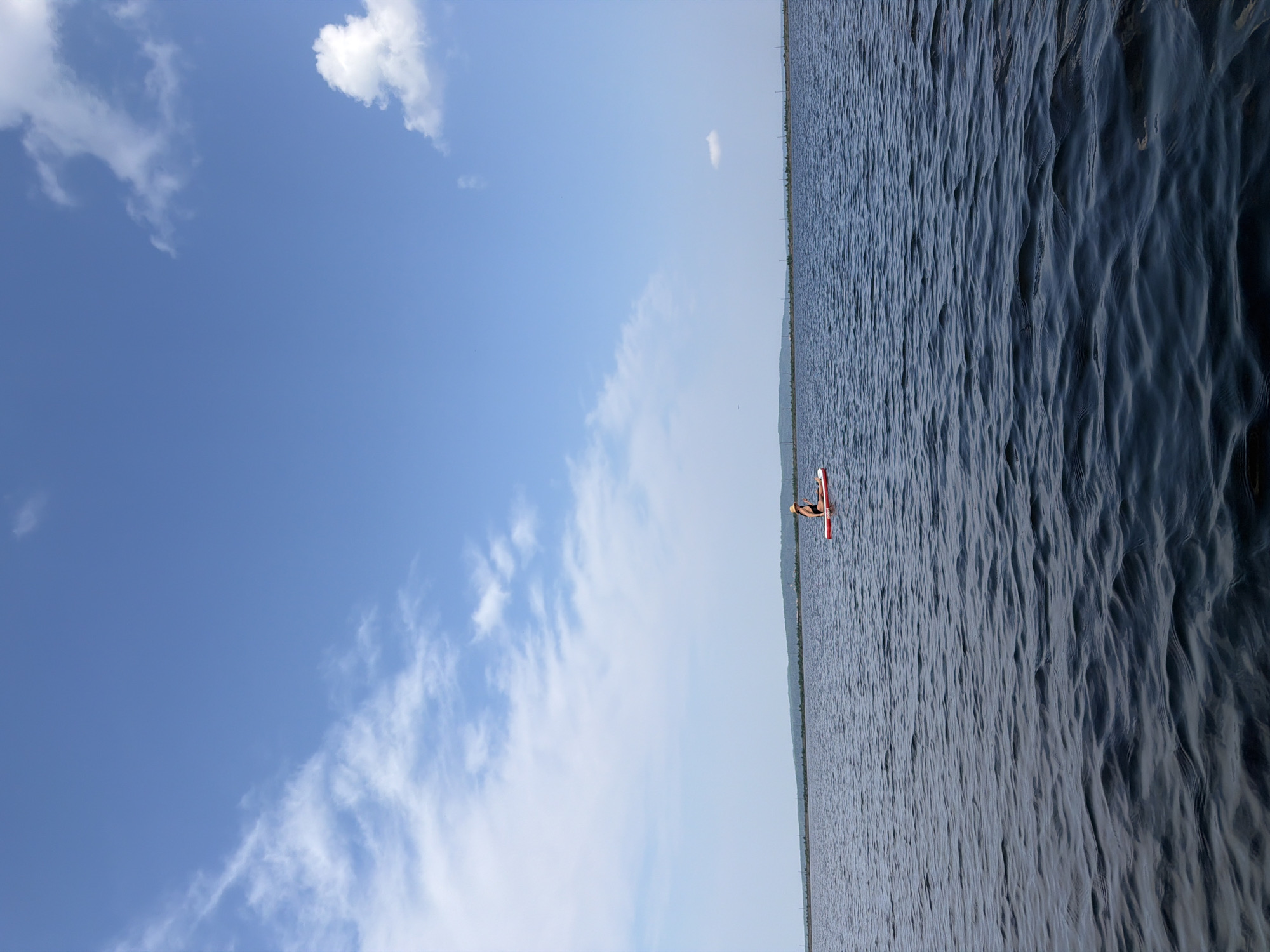 A person wearing a hat and a swimsuit is paddleboarding on a large body of dark blue water. The person is seated on a red paddleboard. The water has moderate ripples. In the background, a long, low, green landmass is visible on the horizon under a blue sky with scattered white clouds. Power lines or thin, tall structures are dotted along the horizon line. The scene appears to be a natural or semi-natural outdoor environment. The provided location information (Chaikovsky, Russia) does not contain any discernible visual cues in the image itself.