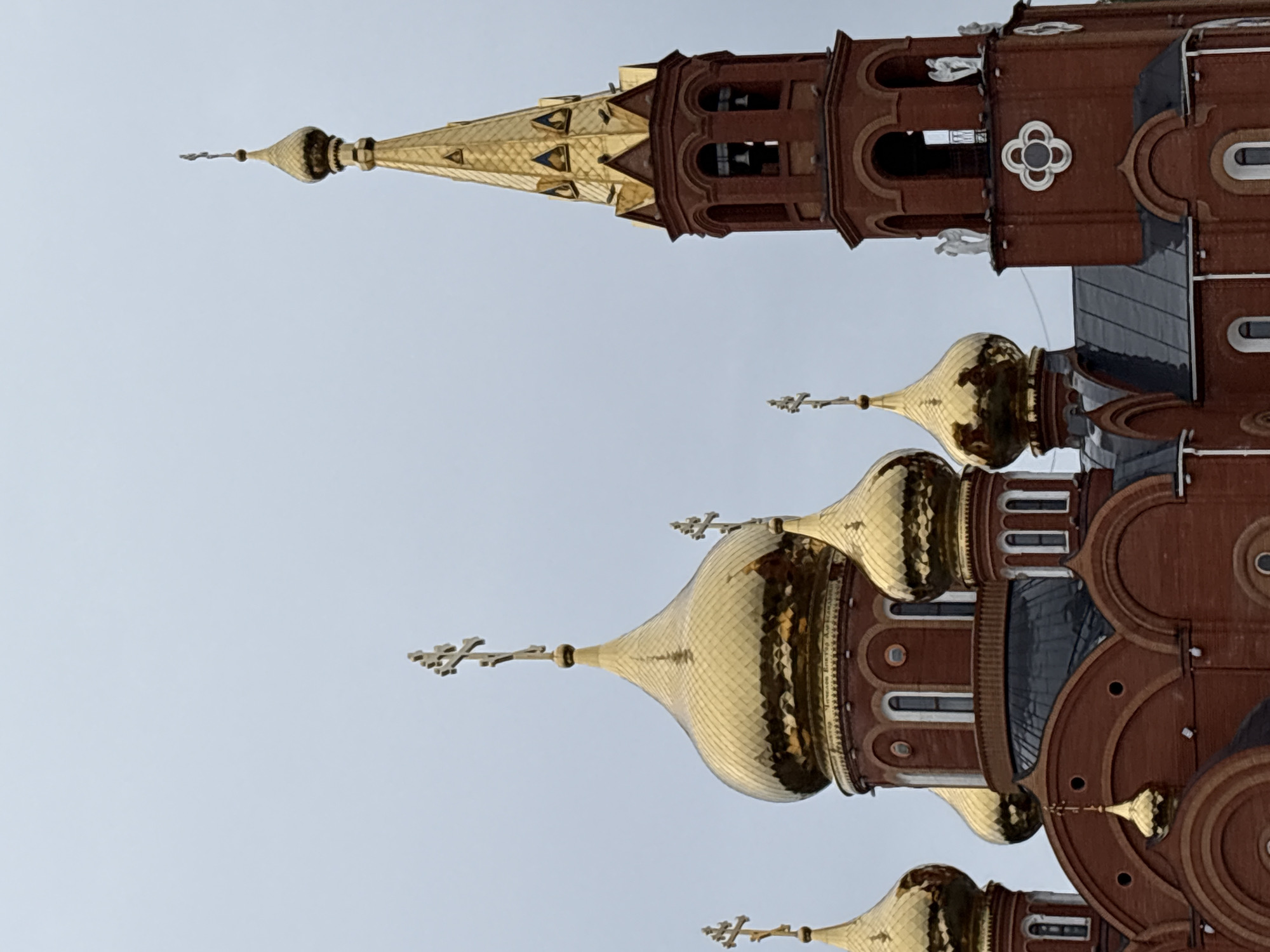 The image depicts a section of the Holy Great Martyr George the Victorious Cathedral in Chaikovsky, Russia. The structure is characterized by red brick and gold domes. The domes are topped with Orthodox crosses. A tower with a gilded, pointed roof and a bell tower are visible. The background is a pale, overcast sky.