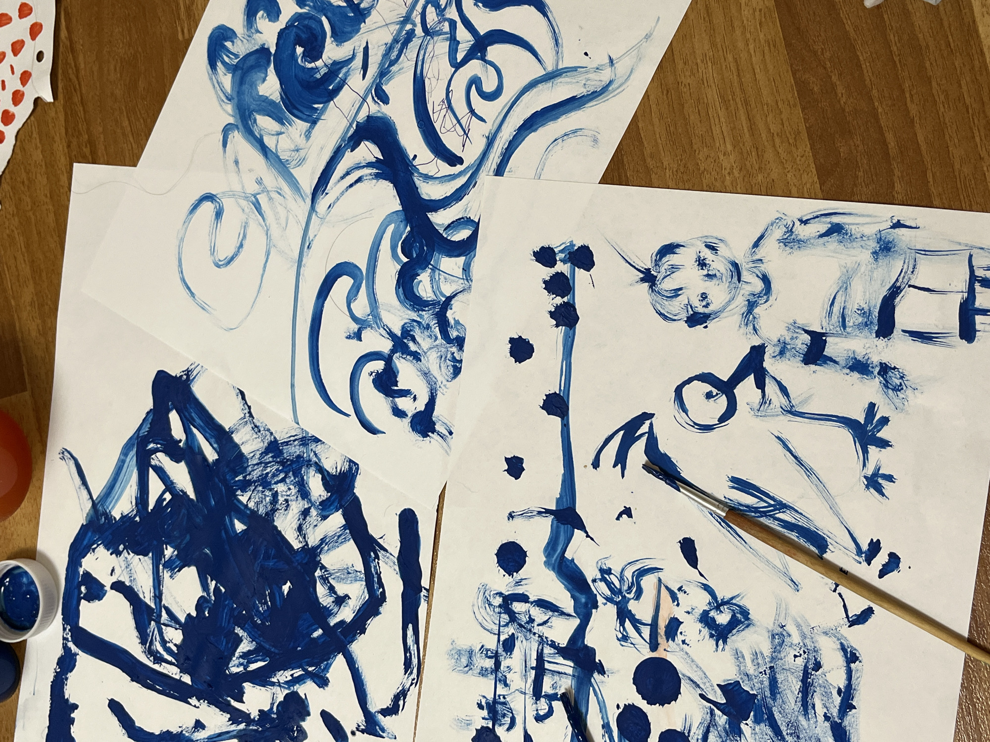 A top-down view captures an art activity in progress, featuring several pieces of white paper covered in blue paint on a wooden surface in Chaikovsky, Russia. The scene is dominated by expressive, abstract blue paint strokes and designs, suggesting a spontaneous and creative endeavor.
Three main sheets of paper are visible. The top paper displays flowing, calligraphic blue lines and swirls, with some thinner, darker lines interwoven. The bottom-left sheet is densely covered with broad, energetic blue brushstrokes, creating a more abstract and intense composition. The largest sheet, on the right, features a mix of abstract and rudimentary figurative elements. On its upper right, a simple, round face with eyes and a nose is discernible, next to some loose body shapes. Below this, a vertical sequence of distinct blue dots and other abstract linear forms runs down the center. The bottom section of this paper shows more varied strokes, including some circular dabs of paint. A wooden-handled paintbrush lies across this paper, indicating its recent use.
Adjacent to the bottom-left paper, a small white pot filled with vibrant blue paint is visible. A red or orange cup is partially seen near the paint pot, and in the top-left corner, a white paper cutout with red polka dots adds another element of craft. The wooden surface, possibly a table or floor, provides a warm, natural backdrop for the vibrant blue artwork. The overall impression is one of creative exploration, likely involving a child given the style of some drawings and the direct application of paint.