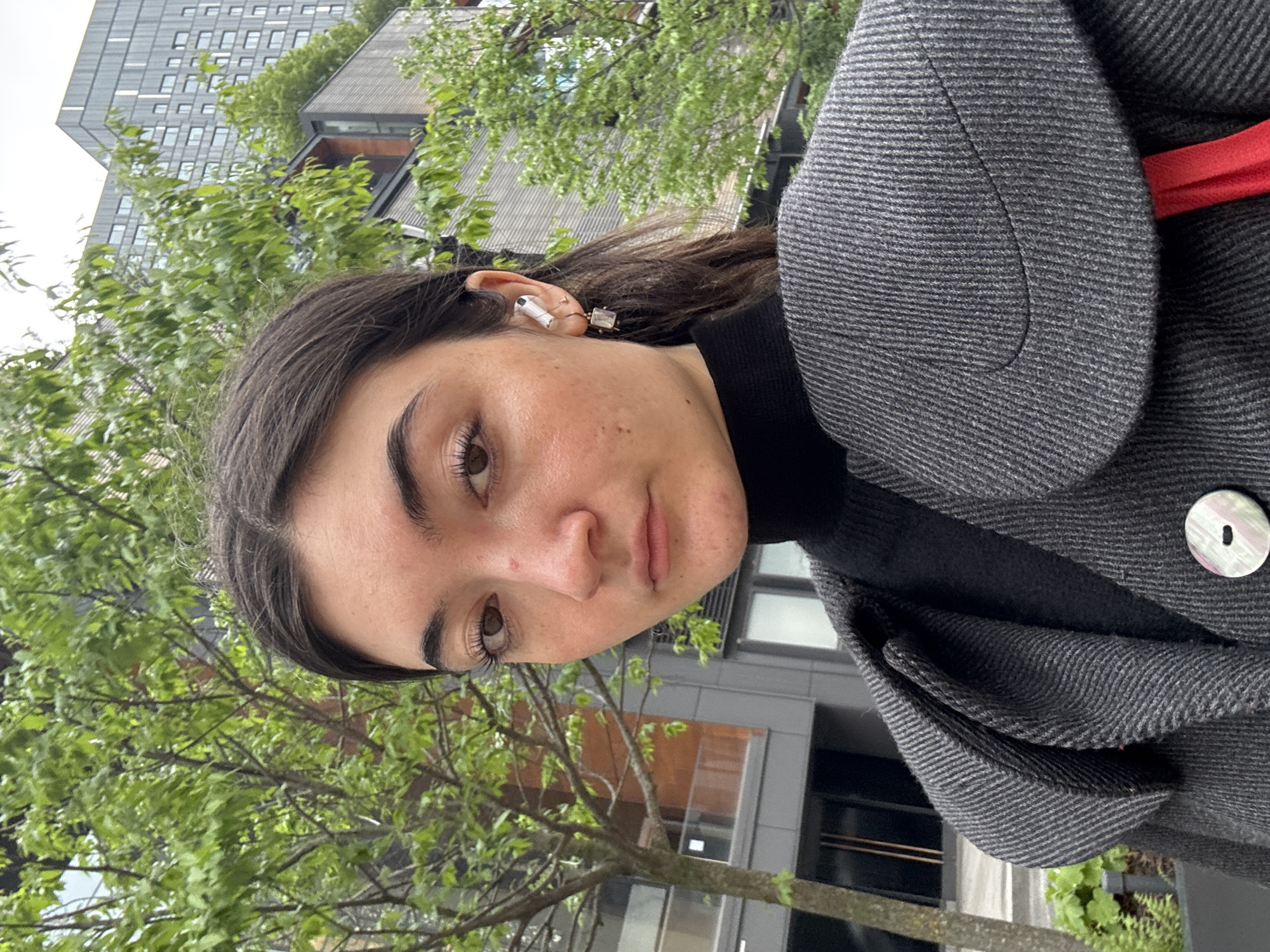 This image is a portrait of a young woman taken outdoors in an urban environment, likely in New York, United States of America. The photograph is oriented vertically, with the subject's head tilted towards the left.
The main subject is a young woman with dark, medium-length hair and prominent eyebrows. Her brown eyes are open, and she looks directly at the camera with a neutral to slightly contemplative expression. Her complexion is natural, showing a few freckles and small blemishes. She is wearing a white, AirPods-style earbud in her left ear, along with a small square earring and a tiny hoop earring on the same ear.
She is dressed in a dark grey coat or jacket with a subtle striped or textured pattern, worn over a black turtleneck. A flash of vibrant red fabric is visible inside the coat's lapel on her right side, and a large, iridescent, mother-of-pearl-like button is prominent on the coat's front.
The setting appears to be a city street or park area during the daytime. The lighting is soft and even, suggesting an overcast sky. In the background, there are lush green trees and modern buildings. One building, particularly visible in the upper left, features a striking contemporary design with numerous square windows and a green, vegetated rooftop. Another building has a dark, angled roof. No discernible text is visible in the image.