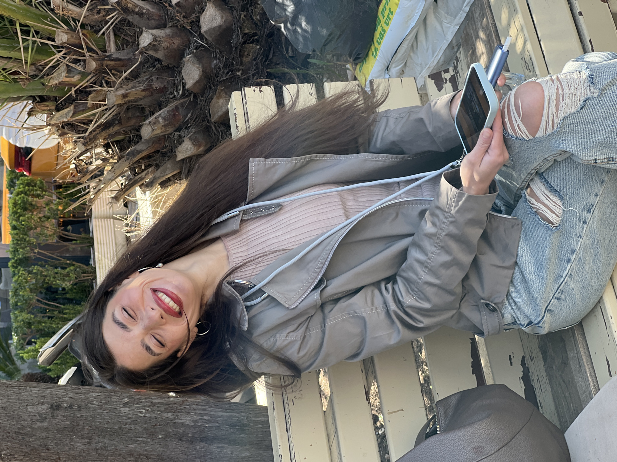 A young woman with long, dark brown hair is sitting on a white, wooden bench in an outdoor setting. She is holding a light blue smartphone and what appears to be an electronic cigarette device. She wears a light grey jacket, a pale pink sweater, and ripped blue jeans. Her location is potentially in Budva, Montenegro. The setting includes a palm tree trunk to the left, as well as foliage and a building in the background. There is a bag behind her on the bench, as well as a black plastic bag and a white and yellow bag.