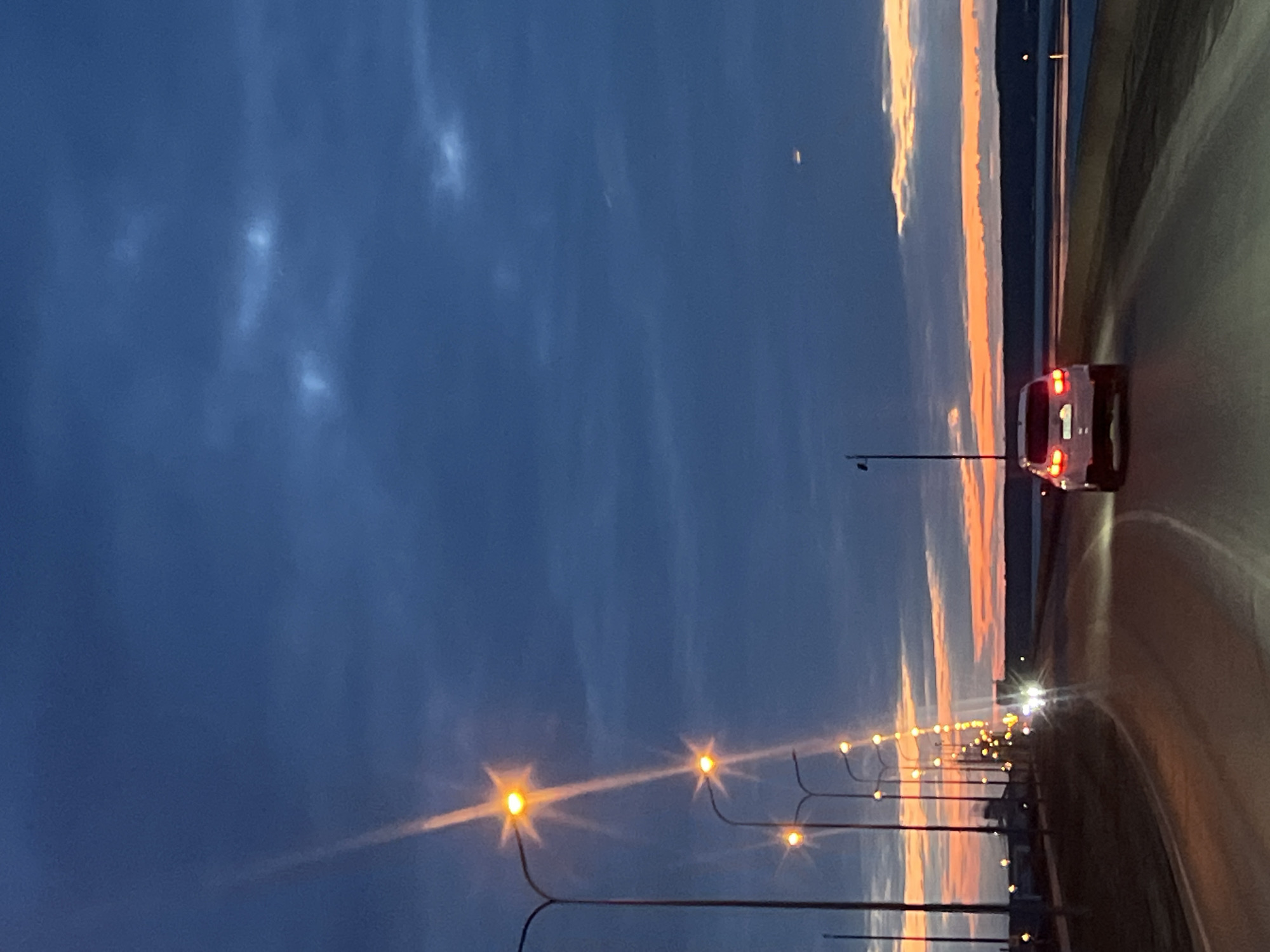 A nighttime photograph taken in what appears to be the city of Chaikovsky, Russia, shows a paved road illuminated by streetlights leading towards a body of water. A white car drives on the road. The sky is a gradient of blues and oranges indicating either sunset or sunrise. Street lights line the side of the road.