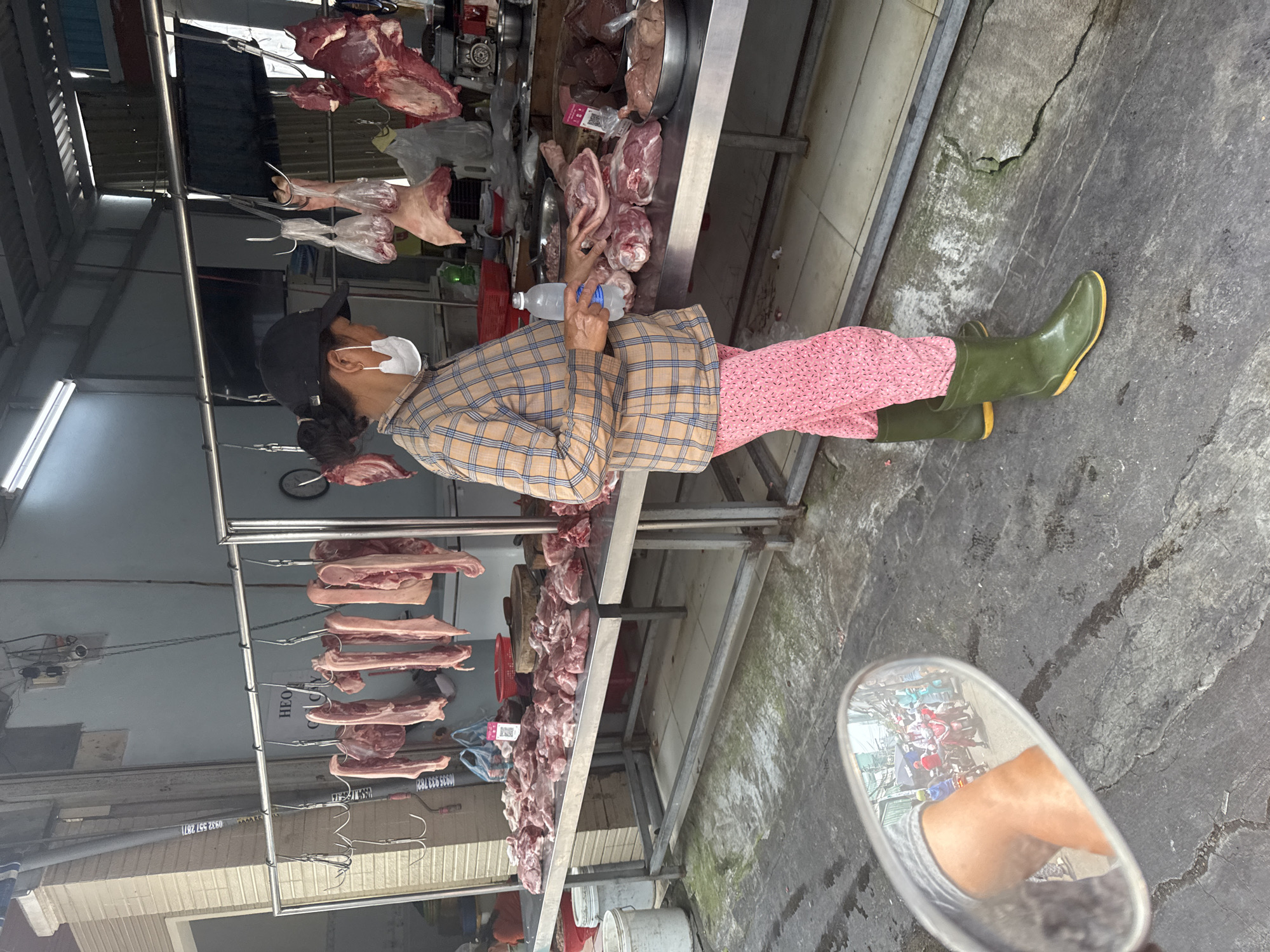 A woman wearing a face mask, a plaid shirt, and pink floral pants is standing at a meat stall in Đà Nẵng, Vietnam. She is wearing green rubber boots and holding a water bottle. Various cuts of meat hang from hooks above the stall and are displayed on the stainless steel counter. The stall is in an open-air market with a corrugated metal roof. A motorcycle mirror is visible in the foreground, reflecting the scene behind the photographer. The ground is concrete.