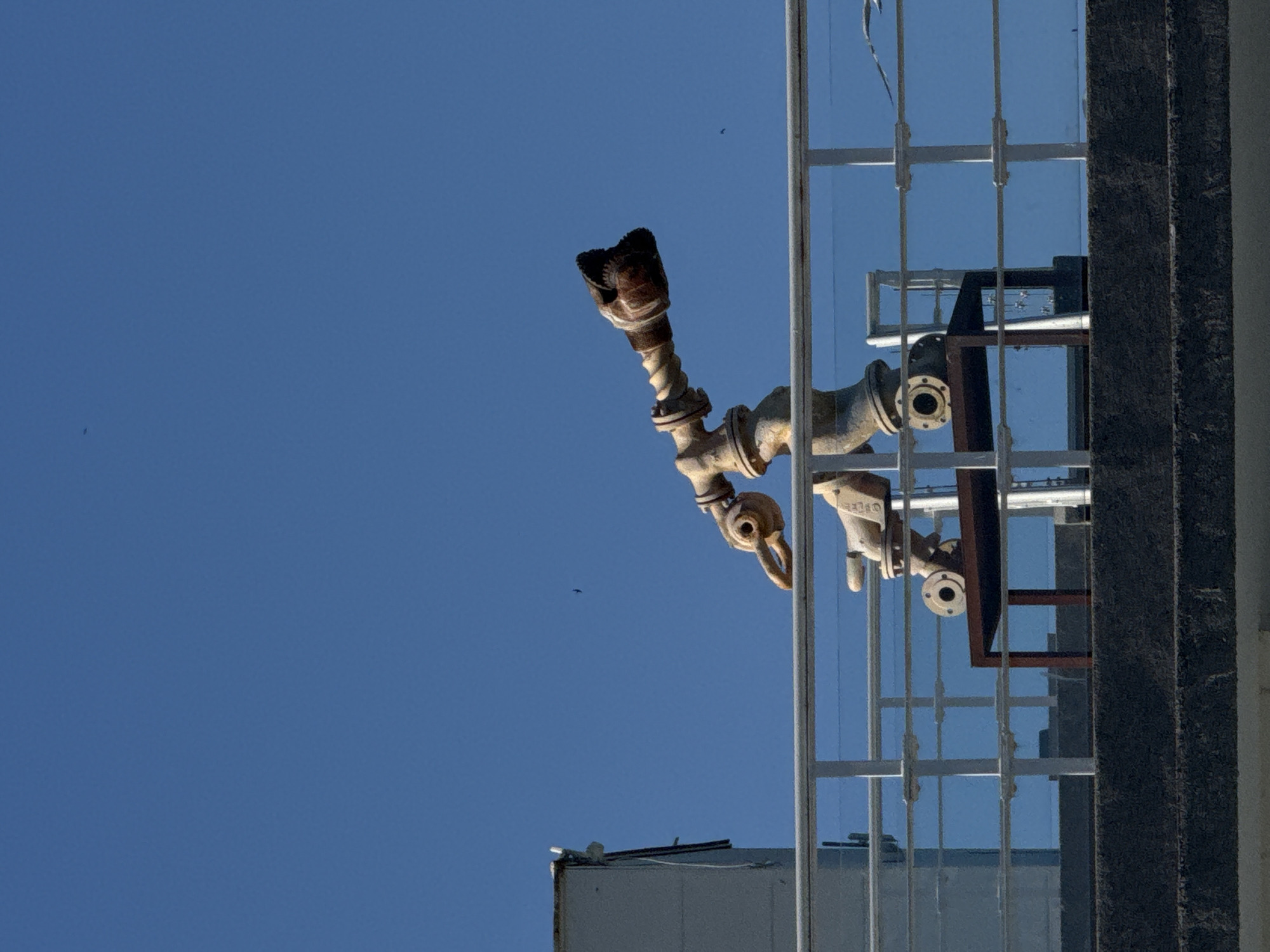 A low-angle shot focuses on an exterior industrial fixture in Burgas, Bulgaria. The fixture is comprised of metal pipes and valves. It is vertically mounted to the exterior of a building, which features a glass panel and metal grid window design. A piece of furniture, partially visible inside the building, reflects the blue sky. The sky is clear. A bird is in flight in the distance. The overall lighting suggests a sunny day.