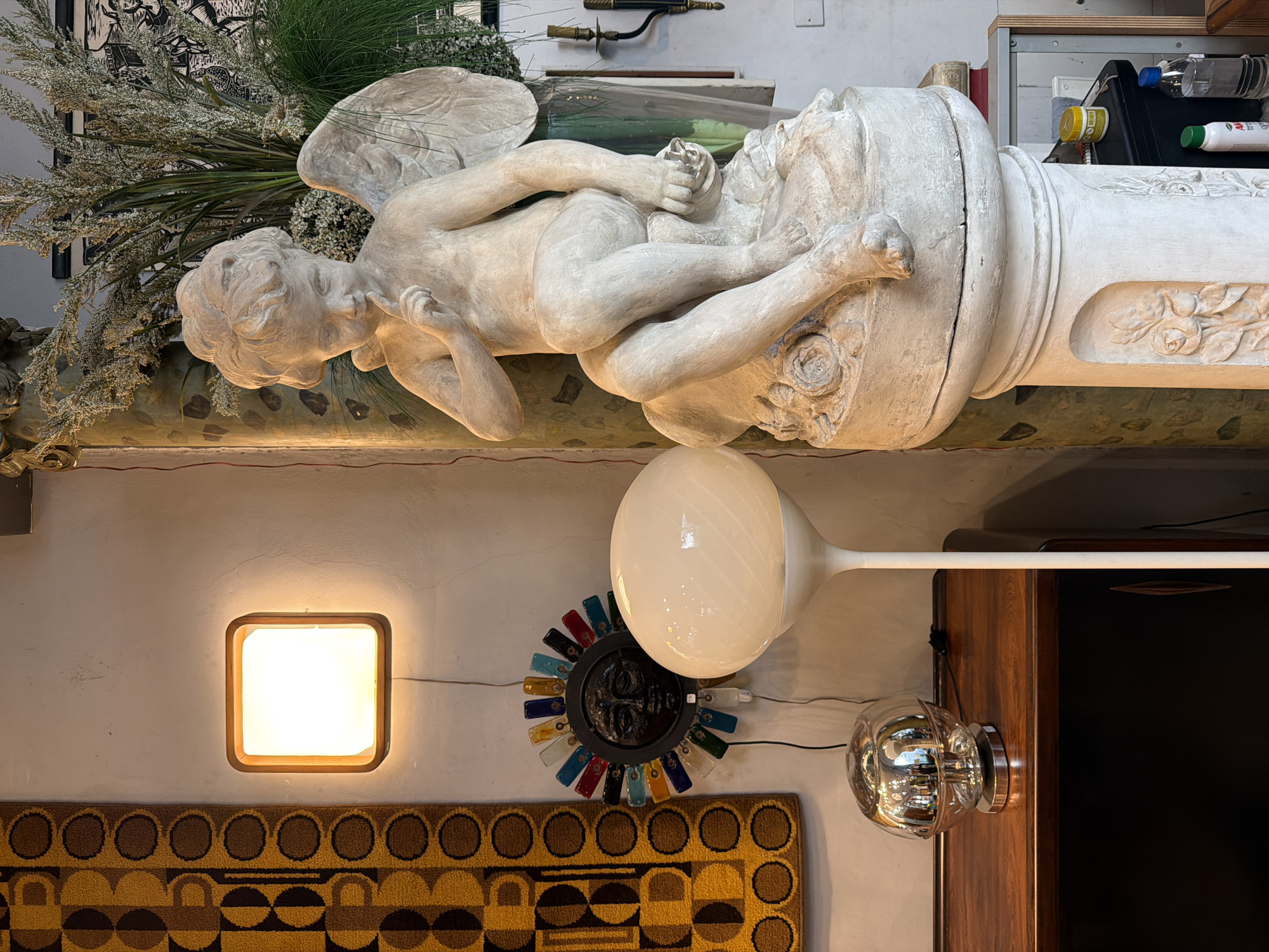 The image shows an interior scene. A pale white angel statue with folded wings rests on top of a decorative column. Next to it are plants and other objects. Below, a square, wooden-framed light fixture illuminates a wall above a patterned rug. A round glass light fixture is mounted on the wall next to a colorful, sun-shaped decoration. A tall, white mushroom lamp is partially visible. A wood and dark paneled cabinet is also present. There is no indication of a location in Buenos Aires, Argentina.