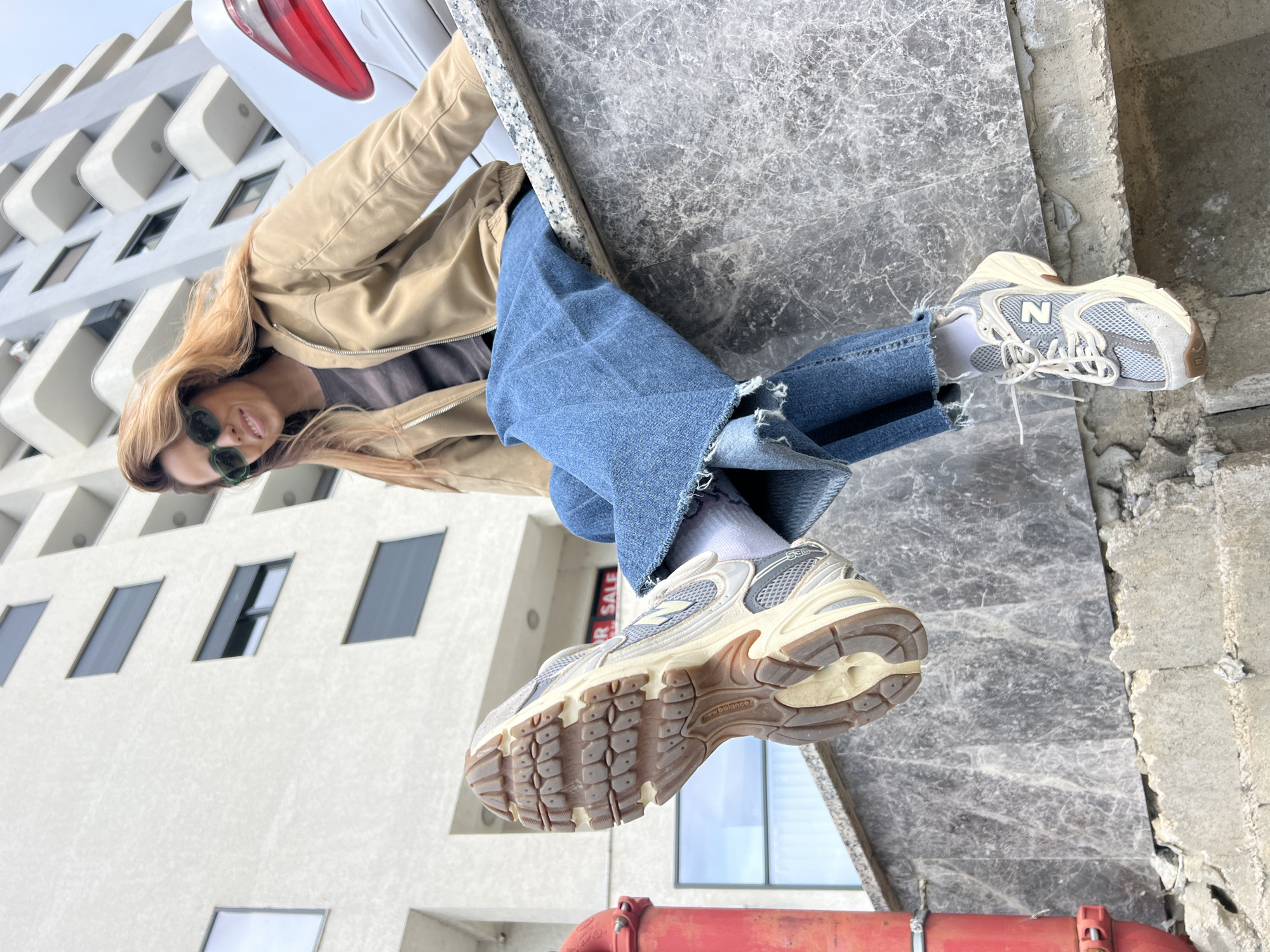 A female subject is depicted seated on a low wall constructed from dark grey veined marble in Bar, Montenegro. She wears a tan zippered jacket, a dark undershirt, wide-leg blue denim jeans with frayed hems, white socks, and light-colored New Balance 530 sneakers. Her right foot is elevated towards the viewer, showcasing the dirty sole of its sneaker. Her left foot hangs below the wall, resting on a rough concrete foundation, with the top of its sneaker visible. The subject has long, light-brown hair, wears green-tinted sunglasses, and is smiling towards the camera. To the far left in the foreground, a weathered red vertical pipe is present. In the background, a modern, multi-story white building with multiple rectangular windows, some equipped with dark roller shutters, and curved balconies is visible. A portion of a white vehicle's rear, including a red taillight, is situated behind the subject on the right. A reflection of a "FOR SALE" sign is discernible in a windowpane behind the elevated right foot.