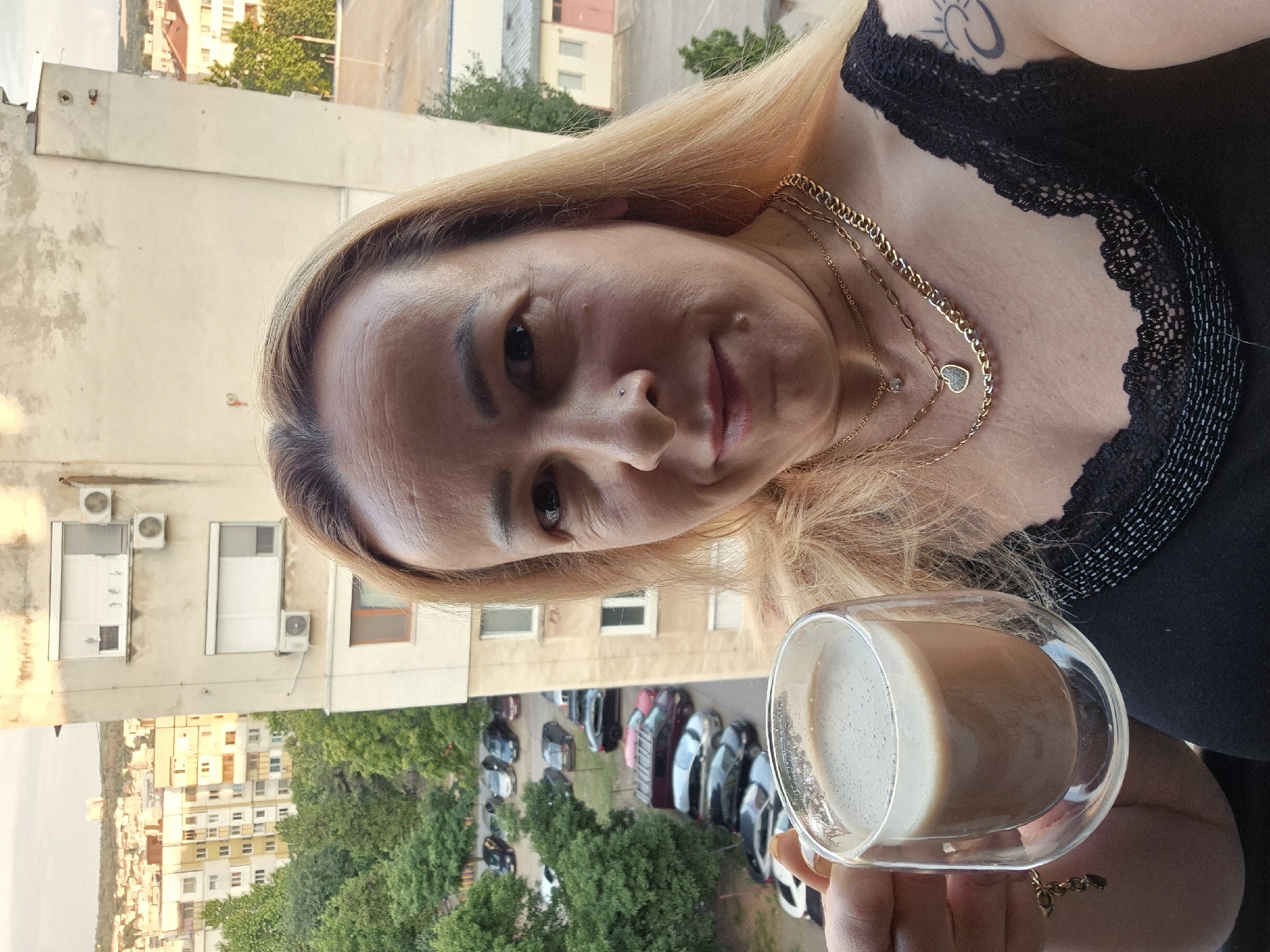 A woman with blonde hair, wearing a black top and multiple gold necklaces, holds a clear glass mug of coffee. She is standing on a balcony, with a city view of buildings and trees in the background. There are several cars parked on a street below. The sky appears to be overcast.