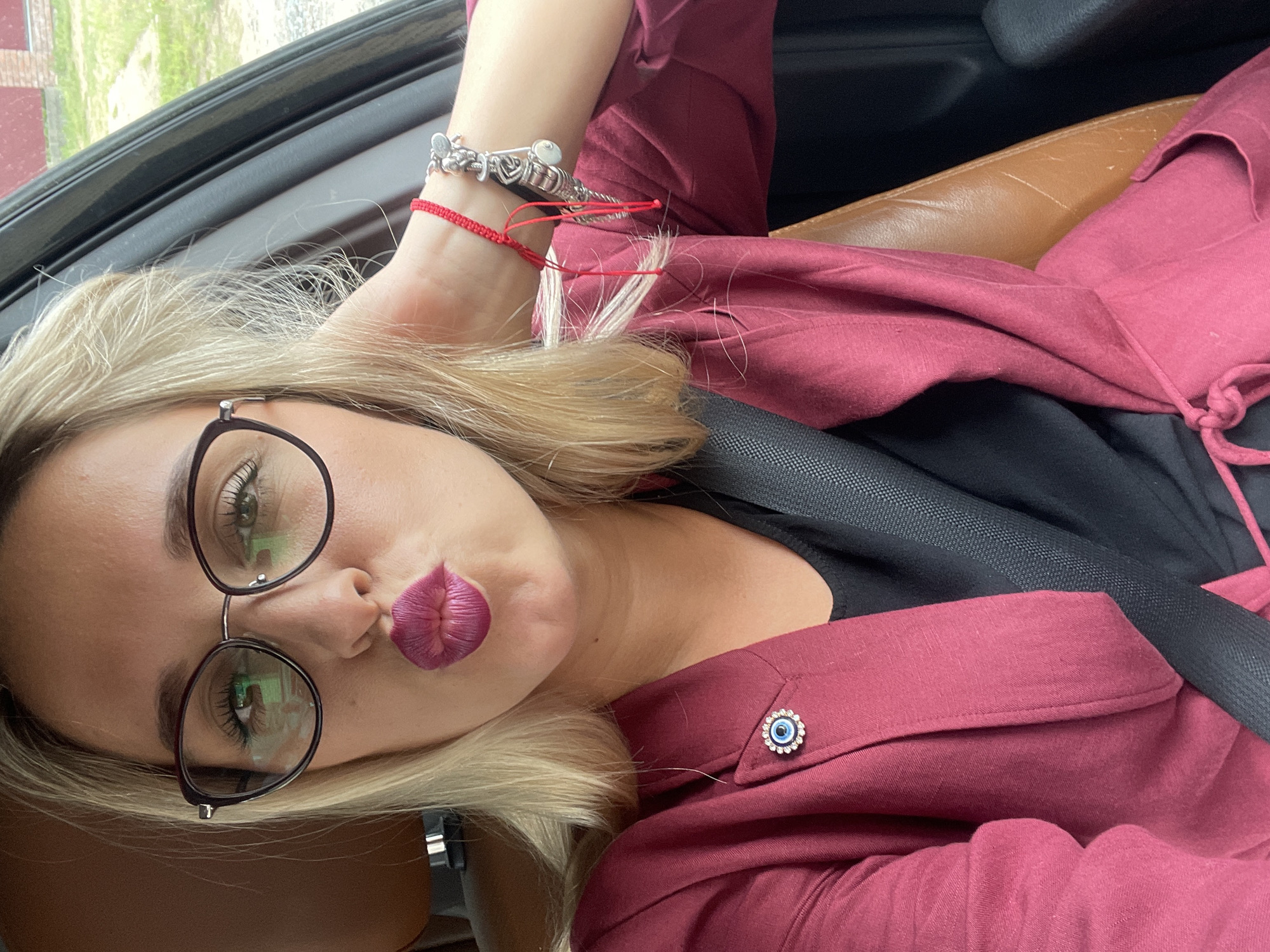A Caucasian woman with blonde hair and glasses is in the driver's seat of a car. She is wearing a maroon blazer over a black top. She is making a duck face and has her hand behind her head, touching her hair. A red string bracelet with charms is on her wrist. The car's interior, including a leather seat and seatbelt, is visible. The exterior scene, seen through the car window, appears to be a rural or suburban area with greenery and a building in the distance. There is no indication of the specific location in Chaikovsky, Russia.