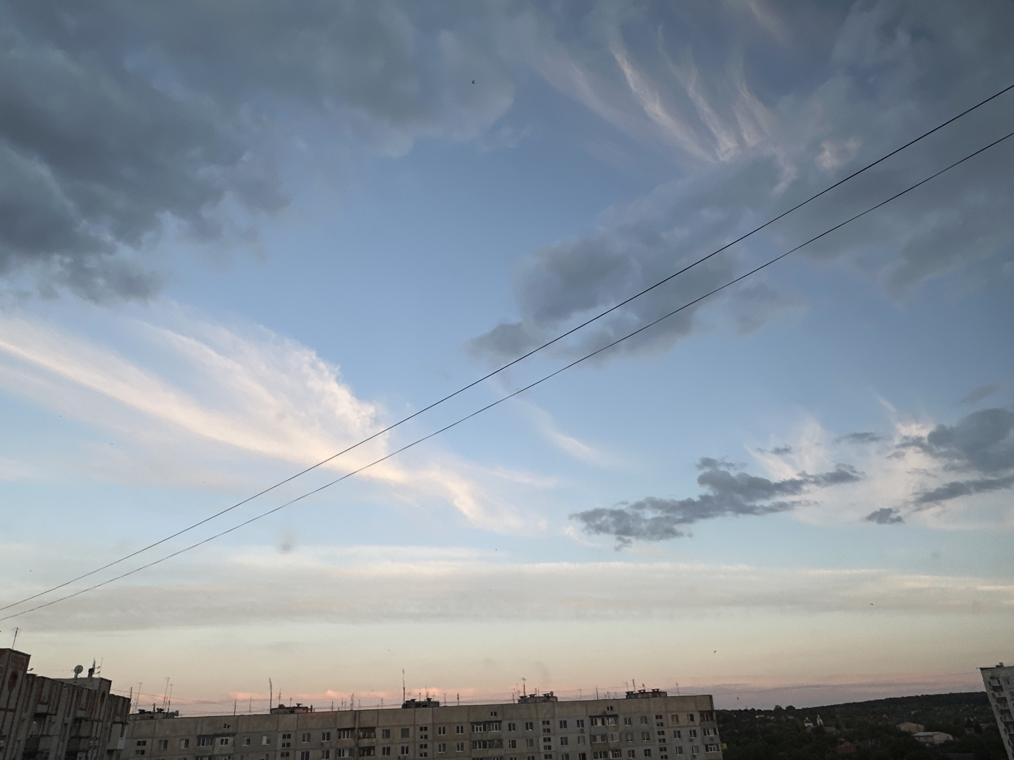 The image depicts a cloudy sky over a city. The sky is a mix of blue and pastel colors, with wispy clouds in the upper portion and darker, more textured clouds towards the sides. Two power lines stretch diagonally across the frame. Below the sky, the rooftops of several multi-story apartment buildings are visible. These buildings appear to be made of concrete and have a somewhat utilitarian design, with numerous windows and some visible antennas or satellite dishes on the roofs. In the distance, a line of trees forms a darker horizon against the lighter sky. There are no people visible in the image. The scene suggests an urban environment, likely during the daytime or early evening, given the lighting conditions and cloud formations.