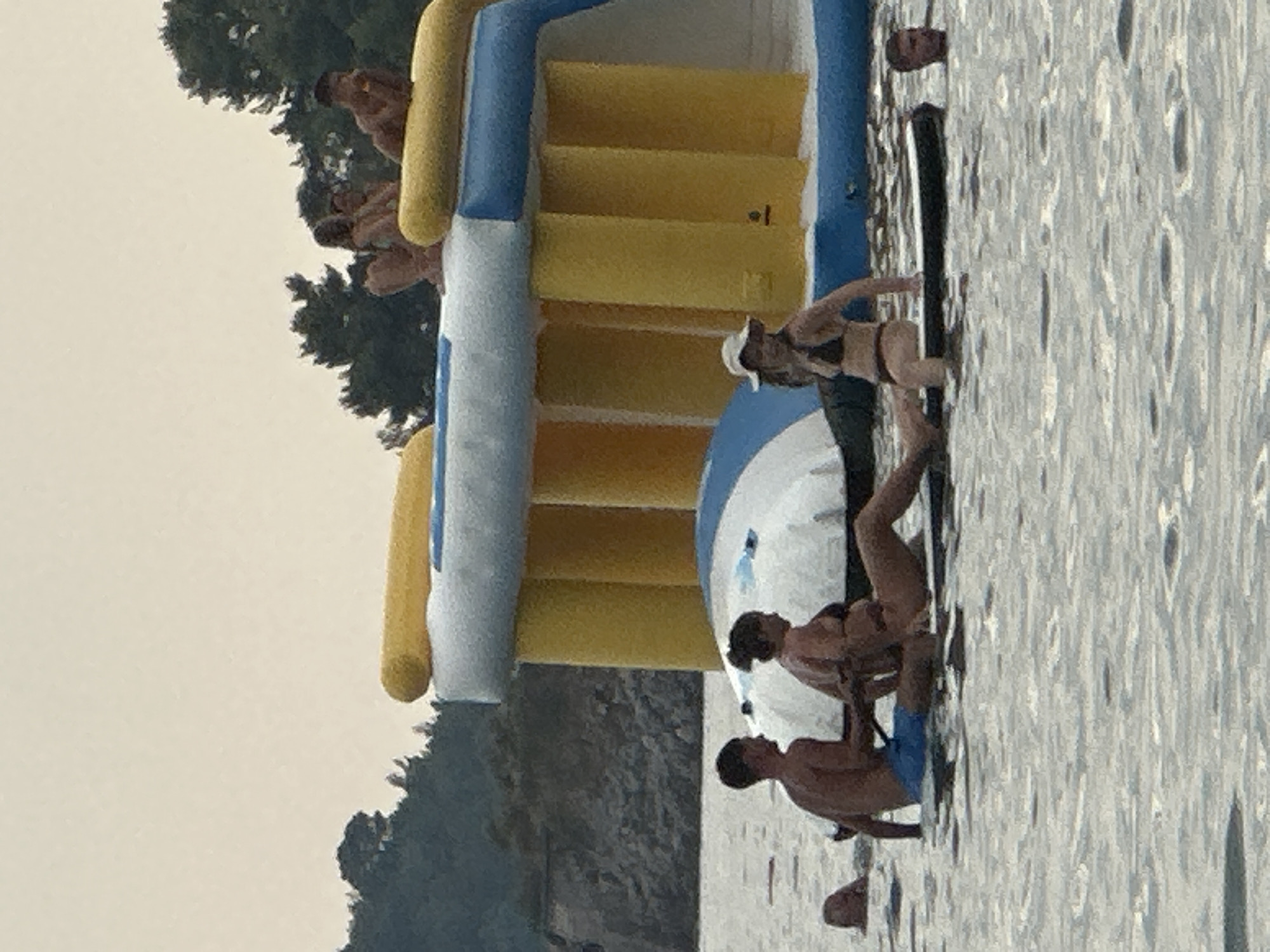 In the waters of Boreti, Montenegro, a group of individuals are enjoying an inflatable water park. In the foreground, two people are seated on a black board in the water, appearing to be on a paddleboard. A man, wearing blue swim trunks, is seated behind a woman who is wearing a bikini. To the right of the paddleboard, a woman wearing a white hat and a bikini is standing in the water. Further back, a large inflatable water structure, primarily yellow and blue, is partially visible. Several other individuals are on the water park structure in the background. The water is calm, and the sky is overcast.
