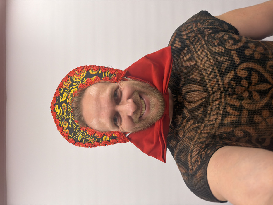 An indoor selfie photograph, taken in Saint Petersburg, Russia, depicts an adult male from the chest up against a plain light-colored wall with a thin red horizontal line at the top. The man, with light brown hair and a blondish-brown beard, is smiling broadly at the camera, displaying his teeth. He wears a black kokoshnik-style headpiece intricately decorated with red and yellow floral patterns and a red scalloped trim. A bright red fabric element, possibly a large bow or gathered collar, is positioned around his neck, resting under his chin. His torso is clad in a short-sleeved, dark brown and light brown/tan patterned top, which exhibits a textured fabric.