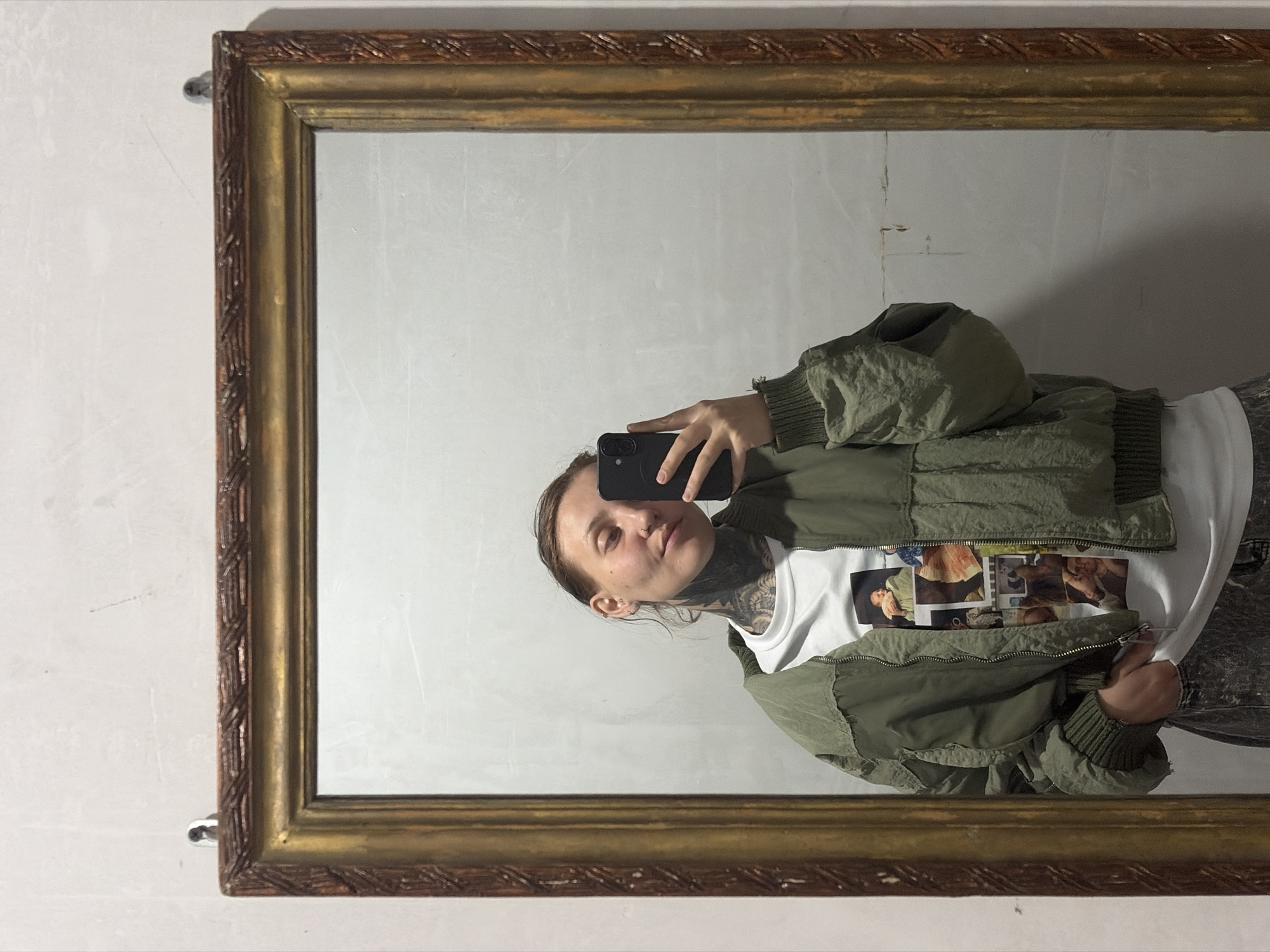 A person is taking a mirror selfie in what appears to be a dimly lit room. The person is standing in front of a large, ornate, gilded mirror and is holding a smartphone in their right hand to capture the reflection. They are wearing a green bomber jacket over a white t-shirt with a collage of images printed on it. The person has extensive tattoos on their neck and chest visible through the t-shirt. Their left hand is in their pocket, and they are wearing dark, patterned pants. The background consists of a plain wall behind the mirror, which has a textured, possibly whitewashed finish. The mirror frame is made of wood with an aged, golden finish and decorative carvings. There are two hook-like objects visible at the top edge of the wall, suggesting the mirror is hung. The lighting is low, creating shadows and a somewhat muted color palette. There is no indication of the location being Malyi Bukor, Russia, or any specific city.