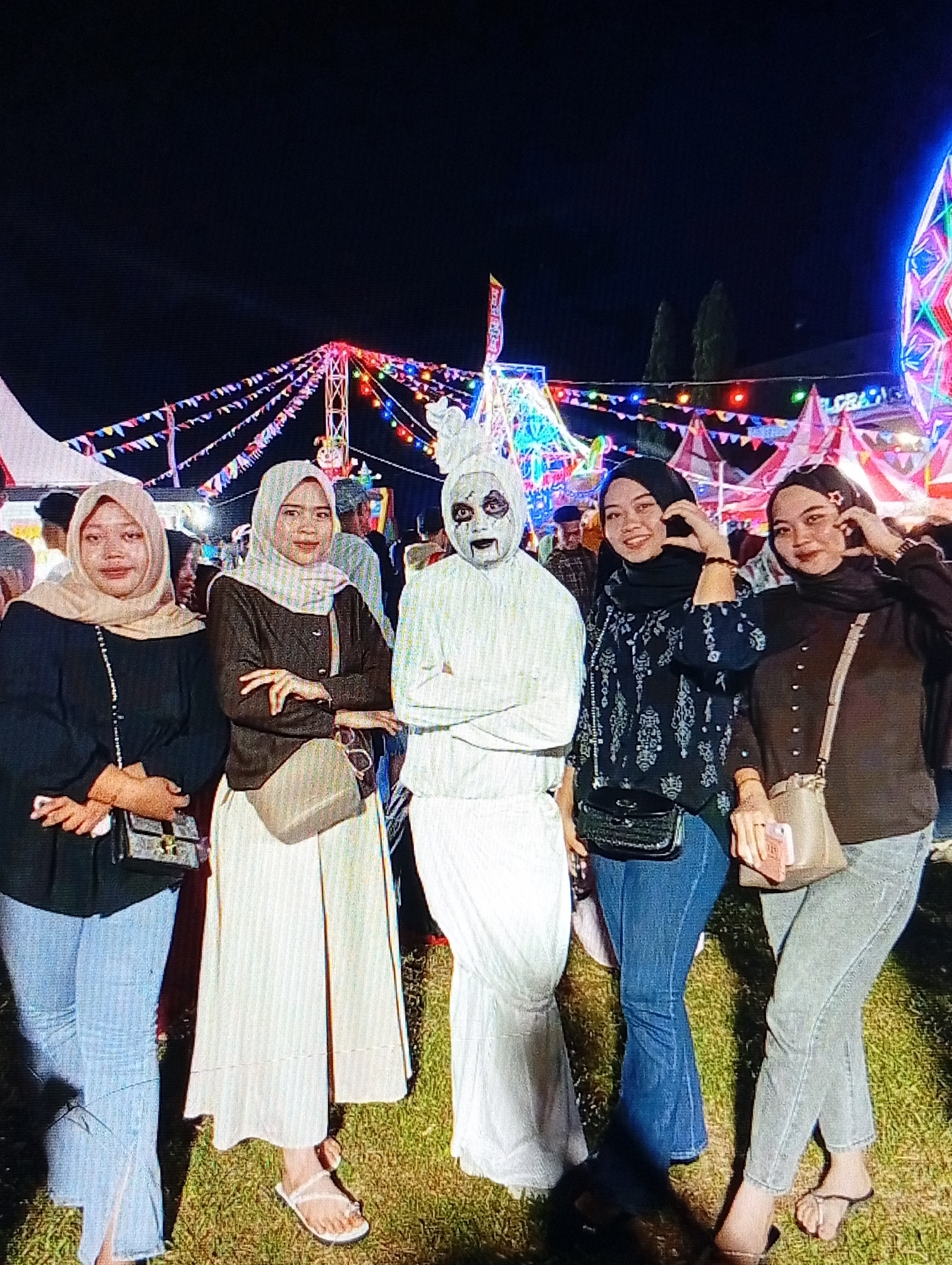 The image depicts an outdoor scene at night, likely at a festival or fairground, given the presence of illuminated structures and colorful decorations resembling strings of lights and flags. In the foreground, five individuals are posed for a photograph. From left to right, the first person is a woman wearing a light brown hijab, a black long-sleeved shirt, and light blue jeans. She carries a black crossbody bag with a gold clasp. The second person is a woman with a light grey hijab, a dark brown long-sleeved shirt, and a cream-colored skirt. She also has a light brown crossbody bag. The third individual is dressed in a white costume that covers their entire body, including their face, which is painted with a white base, black around the eyes and mouth, and red accents, giving them a ghostly or theatrical appearance. The fourth person is a woman wearing a black hijab and a black patterned long-sleeved top with blue jeans. She is holding her hands near her face and has a black quilted crossbody bag. The fifth person, on the far right, is a woman with a black hijab, a dark brown long-sleeved top, and light grey jeans. She has a light brown crossbody bag and is holding a pink object in her right hand. All five individuals are standing on grassy ground. In the background, illuminated structures and more colorful lights suggest a lively event. The context provided indicates this scene is located in the city of Lhokseumawe, Indonesia.