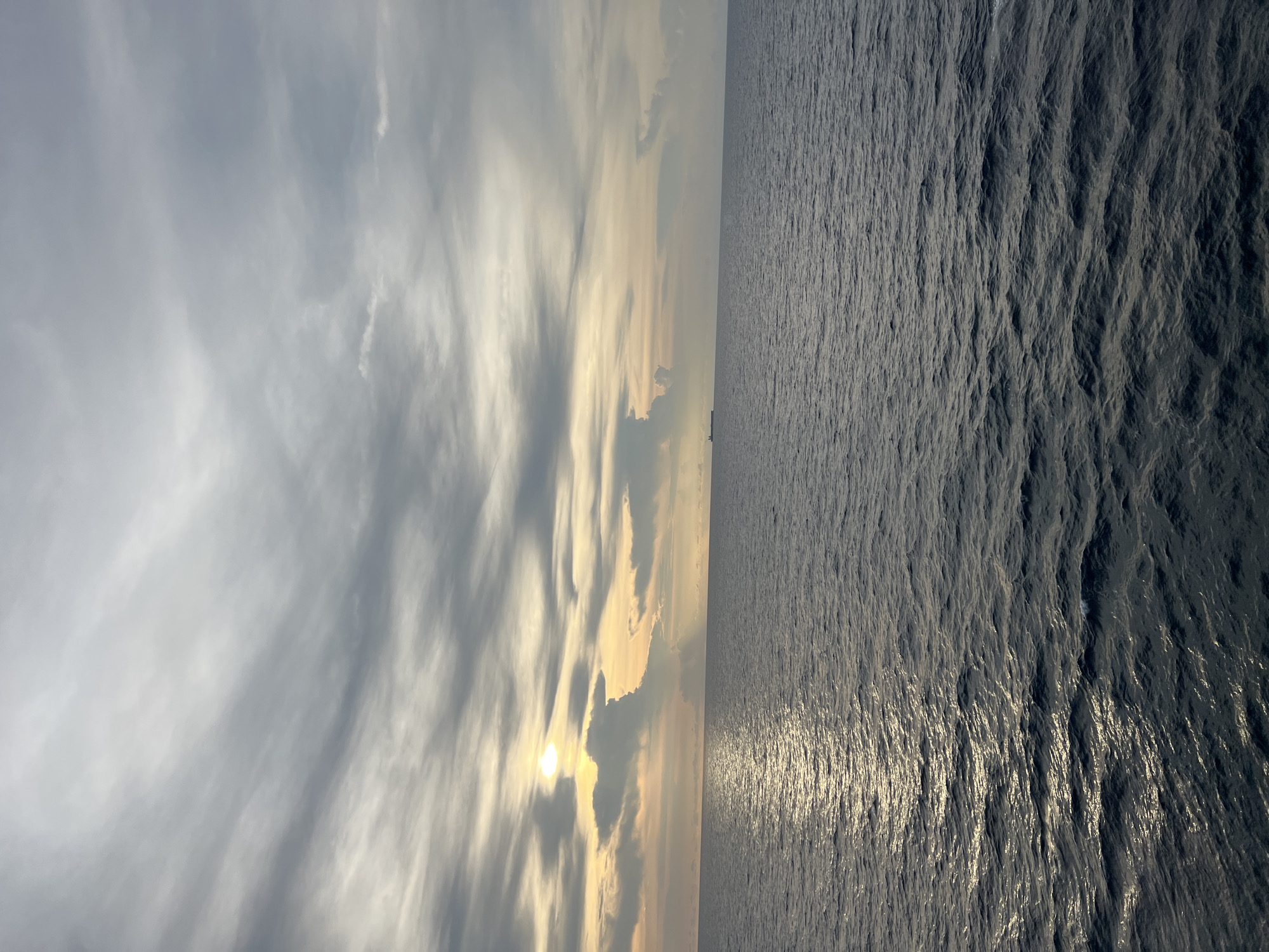 The image depicts a vast expanse of dark, choppy ocean under a cloudy sky. The sun is setting or rising, as indicated by the golden light breaking through the clouds on the left side of the frame and casting a shimmering reflection on the water. There is a distant cargo ship visible on the horizon. The environment is natural and oceanic. There are no people or discernible structures in the image, and no actions are taking place. The location is identified as Unknown, Unknown.