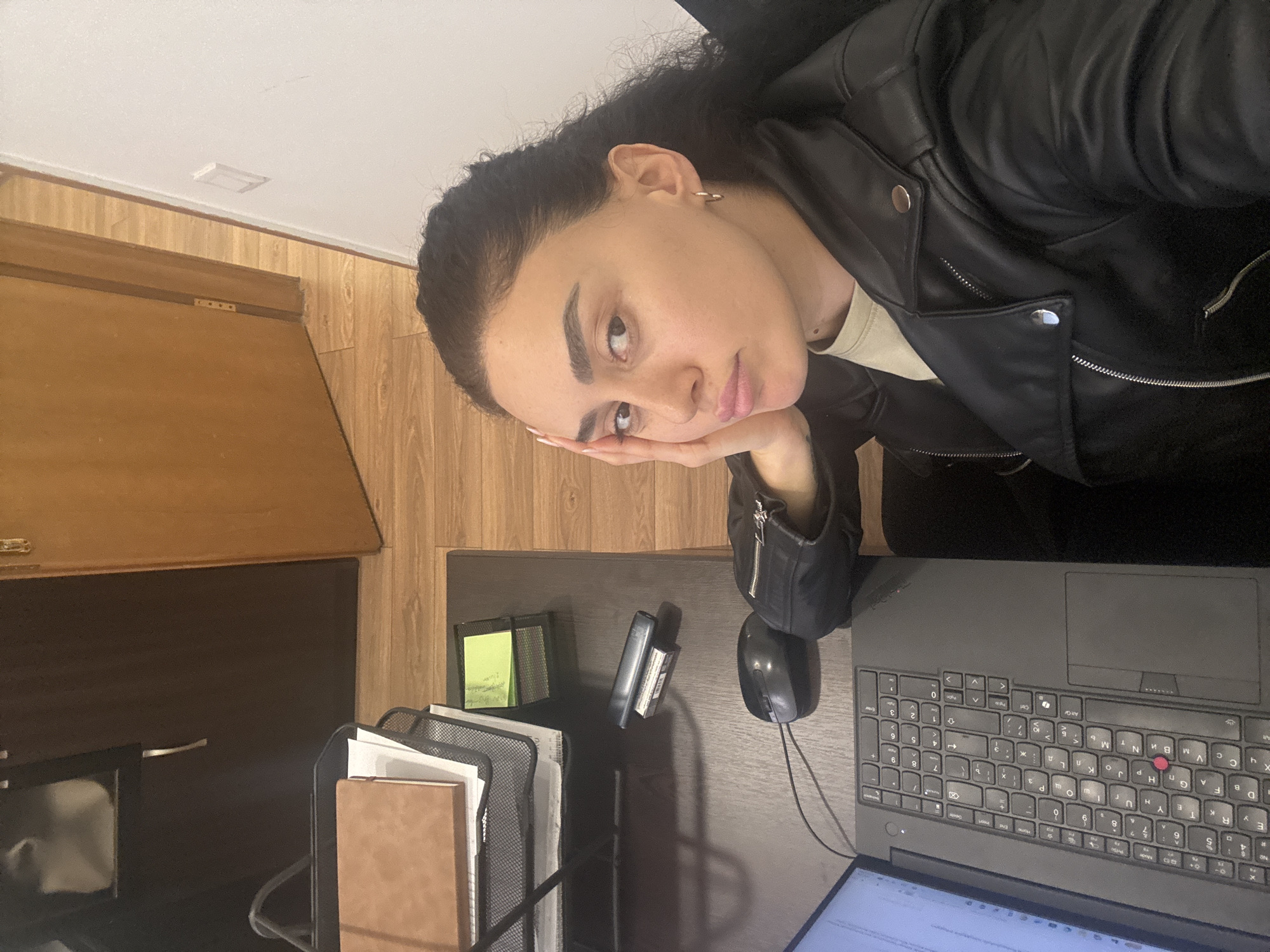 A woman with dark hair is seated at a desk with a laptop, mouse, and documents. She is wearing a black leather jacket and has her hand resting on her face. The desk is situated near a wood-paneled wall and cabinet. The location appears to be an indoor workspace, possibly in Tbilisi, Georgia, but there is no concrete evidence to support that claim.