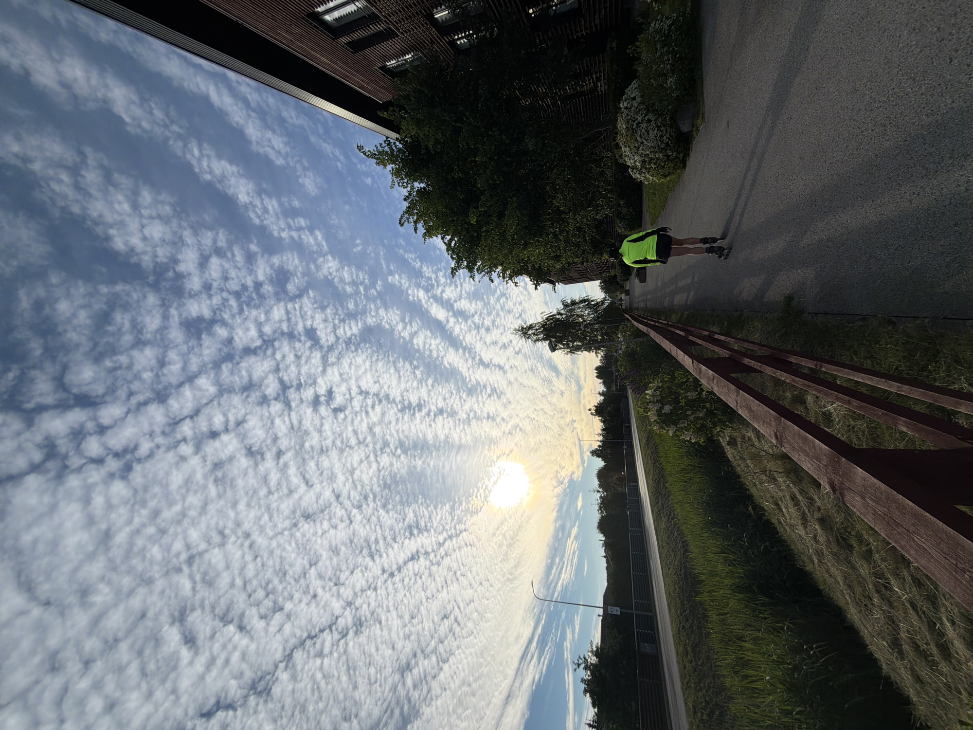 In Tallinn, Estonia, a person on rollerblades is captured from behind, wearing a neon yellow jacket and black shorts, as they move along an asphalt path next to a wooden fence. The path is lined with trees and bushes. To the left of the path, a grassy area slopes down towards a road with streetlights. The sky above is filled with a dramatic pattern of altocumulus clouds, with the sun visible and partially obscured by them. On the right side of the image, a modern brick building with dark horizontal detailing stands, its windows reflecting the sky. The overall lighting suggests it is either early morning or late afternoon.