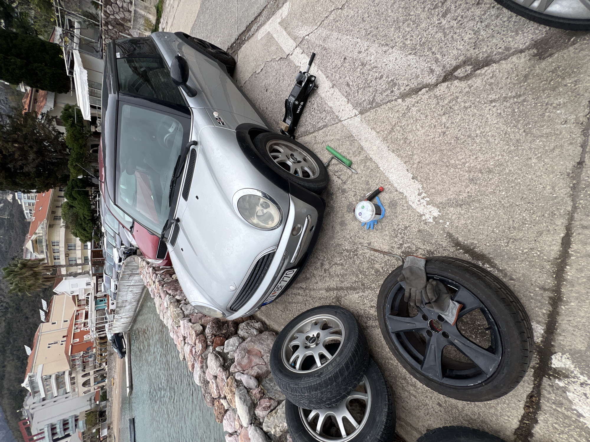 A silver Mini Cooper is parked on a concrete surface next to a stone wall, with its front left wheel removed. The car is lifted with a black hydraulic jack. Three spare wheels are near the wall, two silver and one black. Nearby are gloves, a screwdriver, a lug wrench, and a work light. The location appears to be Rafailovići, Montenegro, based on the architecture and waterfront setting in the background. Other parked cars are visible along the waterfront. The day is overcast.