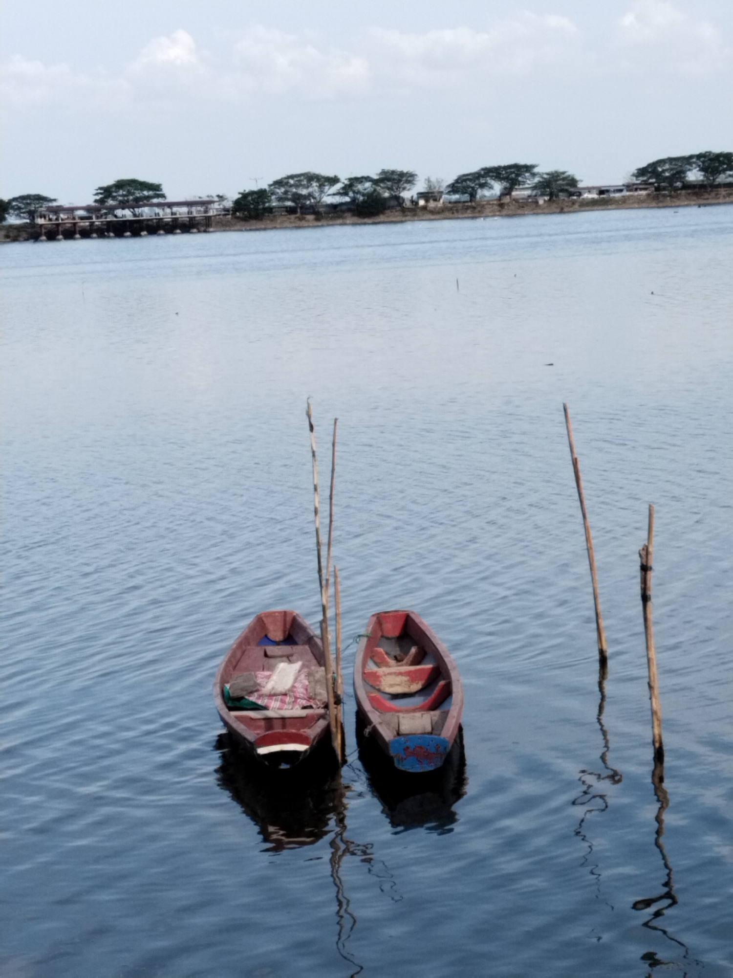 The image depicts a tranquil waterscape in Lhokseumawe, Indonesia, featuring two small, traditional wooden boats afloat in the foreground. Both boats are reddish-brown in color, with subtle variations; the one on the left holds a striped fabric or blanket, while the boat on the right has a distinctive blue-painted stern and appears to contain other items, possibly gear. Tall, slender wooden poles are positioned upright in the water beside the boats, likely acting as mooring posts or markers, and their reflections are visible on the water's surface.
The water itself is calm with gentle ripples, reflecting the boats and poles. In the background, a distant shoreline stretches across the horizon, densely lined with mature trees featuring broad canopies. Behind these trees, a long, low-lying structure, possibly a bridge, dam, or pier, can be seen with multiple supporting pillars extending into the water. The sky above is bright and partly cloudy, suggesting a daytime scene under soft, diffused light. The overall mood of the image is one of stillness and serenity. No individuals are visible, and there is no discernible text in the scene.