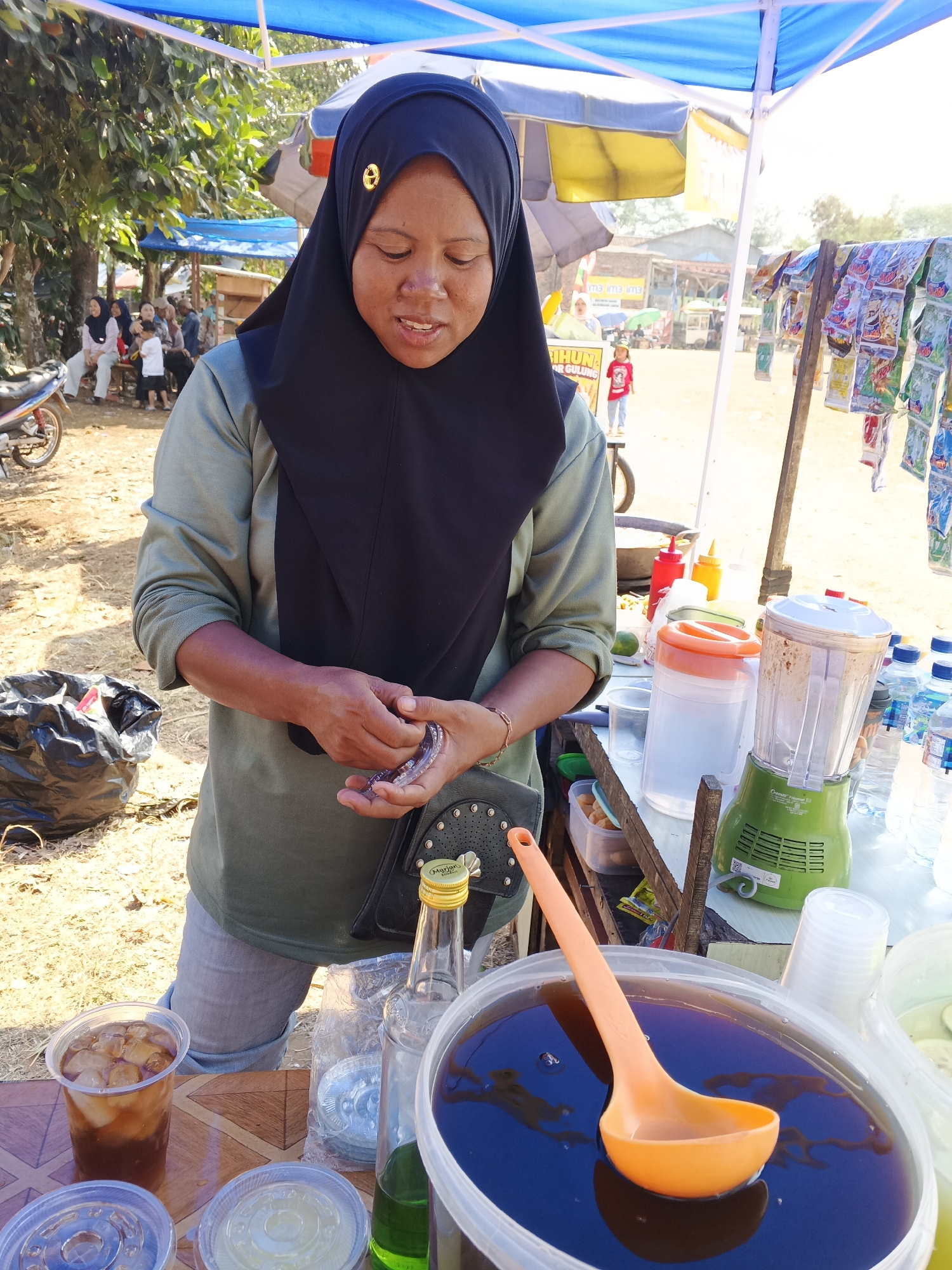 In Warungkiara, Indonesia, a woman wearing a black hijab and an olive green shirt stands behind a market stall. She is holding a small object, possibly a piece of food, in her hands and looking down at it. The stall is set up outdoors under a blue and yellow canopy, with trees and other market stalls visible in the background. There are various items on the stall, including a blender, containers of liquid, bottles of water, and a large bucket filled with a dark brown liquid and an orange ladle. In the foreground, there are plastic cups with drinks and lids, and a black trash bag. The ground is covered in dry dirt.