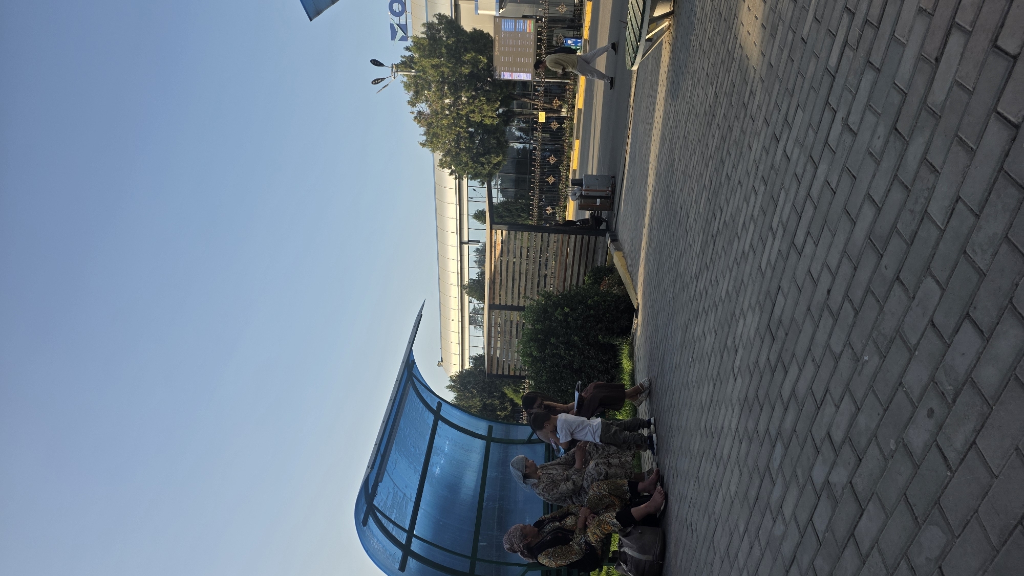 The image depicts an outdoor scene at what appears to be an airport or transportation hub in the city of Nariman, Kyrgyzstan. In the foreground, a paved walkway made of rectangular bricks leads towards the viewer. To the left, a blue translucent awning shelters a bench where three women and a young child are seated. Luggage is visible near the women. The child is standing and looking towards the seated individuals. A second woman is standing behind the seated group.
Further back, a building with a modern facade, featuring large windows and a metallic roof, is visible. Adjacent to the building is a black decorative fence and a wooden slatted structure. To the right of the building, a digital display board with flight information is mounted on a pole. A man in light-colored clothing is walking across the paved area in front of the building, carrying a bag. Trees and greenery are interspersed throughout the scene. The sky is clear and blue, indicating daytime.