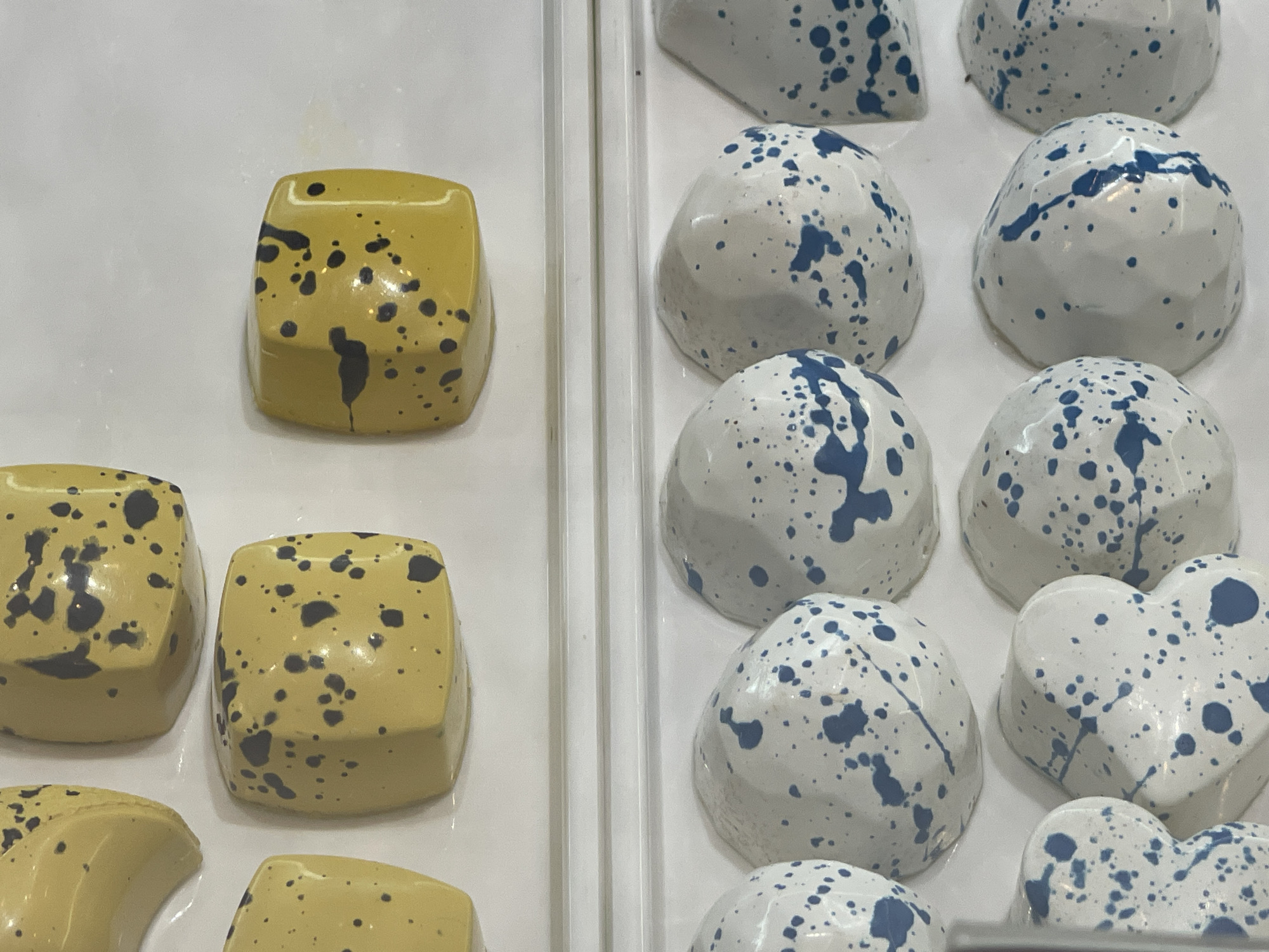 This image shows a close-up of a display of chocolates. On the left side, there are three square-shaped yellow chocolates with dark grey speckles. Below these, partially visible, are two crescent-shaped yellow chocolates with similar speckles. On the right side, there are several white chocolates with blue speckles. These chocolates are shaped like rounded geometric forms, with one distinctly heart-shaped. The chocolates are arranged on a white surface, likely a display tray. The background is a plain white surface. The scene is indoors, and the lighting suggests a commercial setting, possibly a confectionery shop or bakery. There are no people visible in the image. The location is not specified to be in Đà Nẵng, Vietnam.