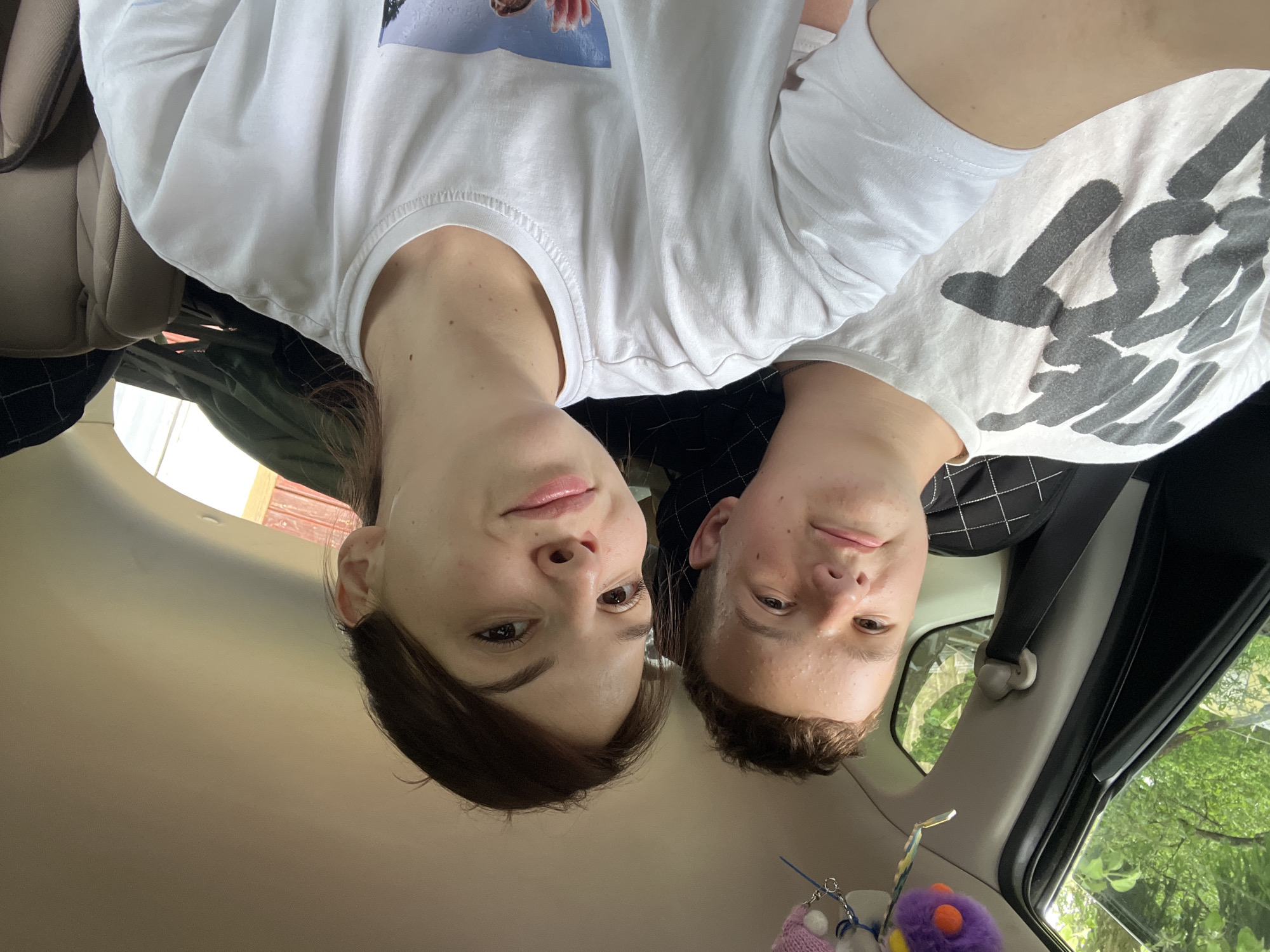 Two young individuals are depicted in a close-up selfie taken inside a vehicle, likely in Мшинская, Russia, during daytime. The male, positioned on the left, appears to be a teenager with short, dark brown hair, a round face displaying visible acne, and a faint smile as he looks towards the camera. He wears a white crew-neck t-shirt with large, dark gray graphic text. The female, positioned on the right and holding the camera, appears slightly older, with dark brown hair and a clear, oval face. She has dark eyes, light pink lips, and a subtle smile, looking directly into the camera. She wears a plain white crew-neck t-shirt with a partial, indistinct graphic visible at the bottom right. A small hoop earring is visible in her left ear.
The car's interior features light beige/gray headliner and pillar trim. The male's seat back shows a dark, possibly black, fabric with a subtle grid pattern, and a black seatbelt buckle and strap are visible. Hanging from the car's overhead area above the male are several small decorative items, including a purple furry toy with orange features and a white knitted object. Through the car windows, lush green foliage and trees are visible, indicating an outdoor, natural environment, with diffuse bright light suggesting an overcast day or shaded area. A dark green object (possibly a bag or clothing) is visible stacked in the rear window area behind the female.