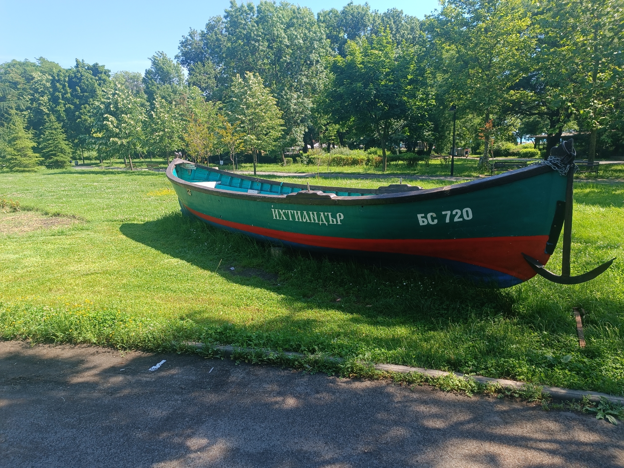 A decommissioned, green-hulled fishing boat with a red waterline and a blue bottom is situated on a grassy embankment in a park. The boat is positioned at an angle, facing towards the right of the frame. The words "ЙХТИАНДЪР" and "БС 720" are visible in white lettering on the side of the boat. An anchor is attached to a chain at the stern of the boat. The background consists of a dense line of green trees under a clear blue sky. The foreground shows a paved pathway casting shadows on the grass. This scene is located in the Sea Garden of Burgas, Bulgaria, a prominent public park situated along the Black Sea coast. There are no people visible in the image.