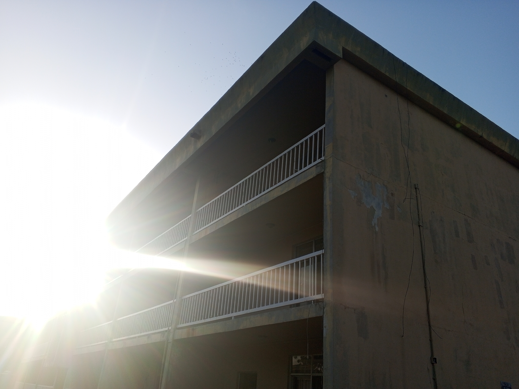 The image shows a three-story building in Tawpo, Nigeria under bright sunlight. The building has a flat roof and open balconies with white railings on the second and third floors. The building's facade is faded and shows signs of weathering and wear. Some electrical wiring is visible on the side of the building. A flock of birds is flying in the sky above the building.