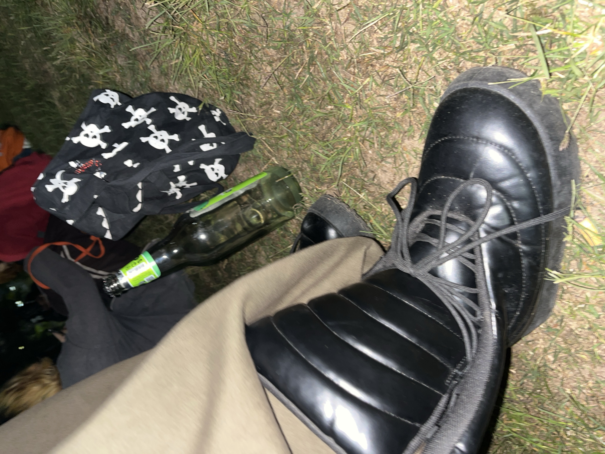A ground-level shot shows a black, treaded boot with untied black laces, a bottle of beer lying on its side, and a black bag patterned with white skulls. The surrounding area is covered with grass. The location appears to be outdoors, but specifics about Buenos Aires, Argentina, cannot be discerned from the image. No people are visible in full. The bottle is a standard beer bottle with a green label. The bag is soft-sided. The boot appears to be on the ground.