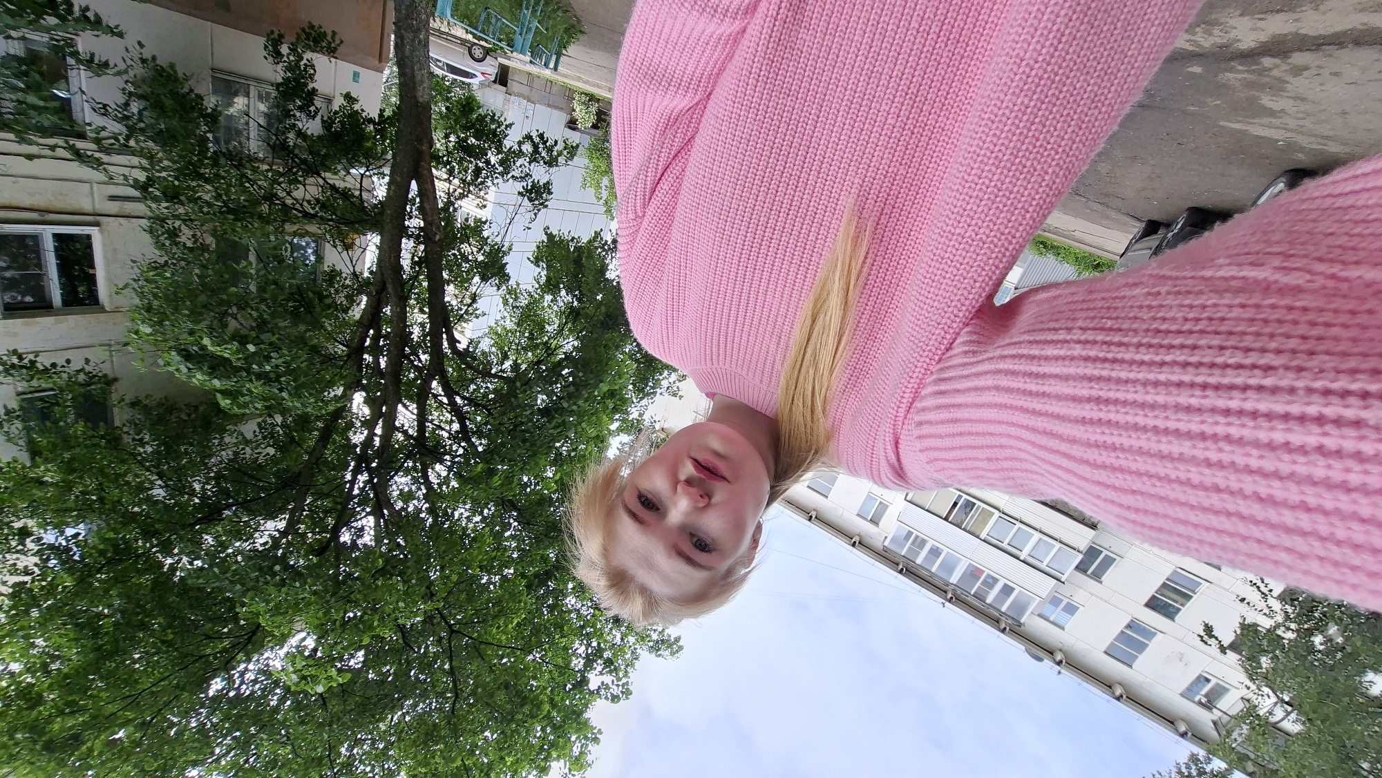 A selfie-style portrait captures a young woman with blonde hair and fair skin, looking directly at the camera with a neutral expression. She is wearing a vibrant pink, ribbed, knitted sweater. Her blonde hair is parted and falls around her shoulders.
The scene is set outdoors, likely in Chaikovsky, Russia, given the architectural style. The perspective is from a low angle, looking upwards. Behind the woman, to her left, is a multi-story residential building with multiple windows and balconies, characteristic of Soviet-era apartment blocks, featuring a light-colored facade. Lush green trees with dense foliage are prominent throughout the background, surrounding the building and extending to the right side of the frame. Patches of clear blue sky with some white clouds are visible between the tree branches and above the buildings. In the lower right portion of the background, a paved surface, possibly a street or sidewalk, and a white car are visible.
The bright lighting suggests it is daytime, and the full greenery on the trees indicates it is either late spring or summer. There is no visible text in the image. The overall impression is a casual outdoor portrait in an urban green environment.