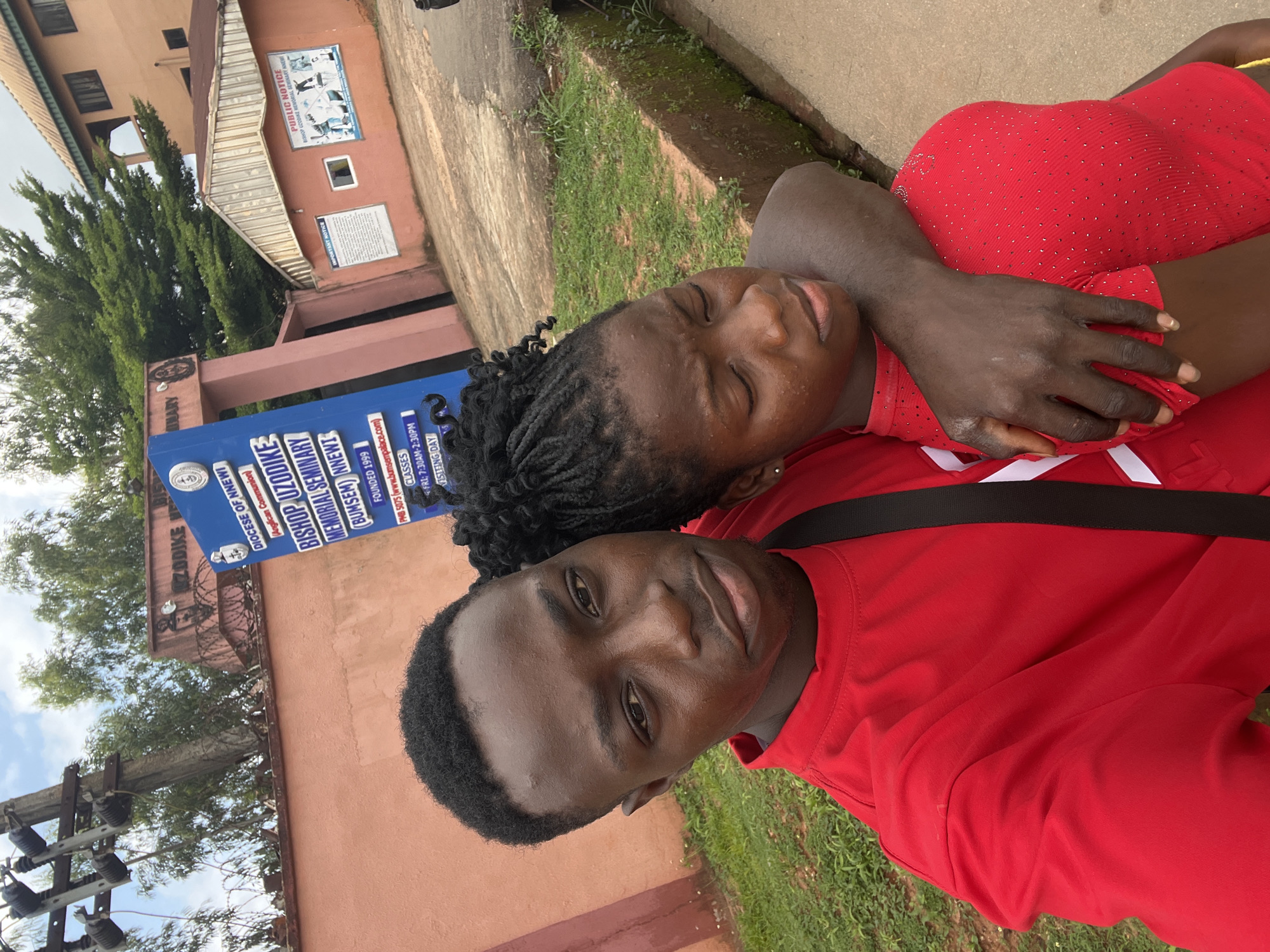 A medium shot captures a young Black couple in a selfie, with the man in the foreground embracing the woman. Both are wearing red tops. The man, on the left, has short, dark hair and is looking directly at the camera with a slight smile. The woman, on the right, has her eyes closed and her head resting on his shoulder, her dark braided hair pulled up. Behind them, a large blue sign with white lettering reads "DIOCESE OF NNEWI" at the top, followed by details about "BISHOP UZODIKE MEMORIAL SEMINARY (BUMSEM) NNEWI." Further down the sign, it states "FOUNDED 1999" and lists "CLASSES FRI: 7:30AM-2:30PM" and "VISITING DAY." The background reveals a salmon-colored building with green foliage and trees. To the right of the sign, a smaller pink building has a "PUBLIC NOTICE" sign featuring images of people in athletic attire. The area appears to be an outdoor urban environment, possibly in the city of Nnewi, Nigeria, given the signage.
