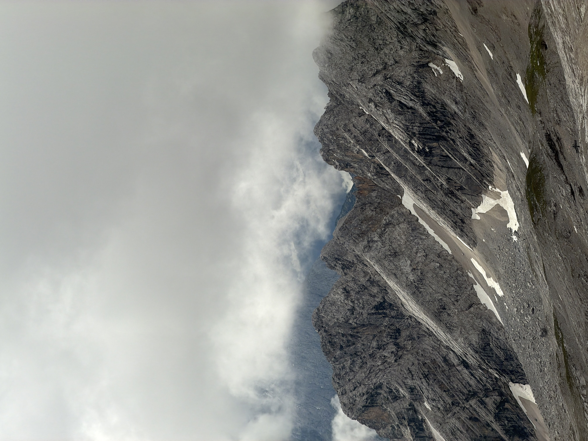 This image depicts a dramatic mountain landscape under a cloudy sky. The foreground is dominated by rugged, gray rock formations with visible striations, suggesting significant geological history. Patches of snow and ice are scattered across the rocky slopes, indicating high altitude and potentially cooler temperatures. The middle ground features more distant mountain peaks, partially obscured by swirling clouds and mist. The sky is filled with heavy, overcast clouds, creating a diffused light that softens the scene. The overall impression is one of wild, untamed nature.
This landscape is characteristic of the Zugspitze massif, the highest peak in Germany, located near Garmisch-Partenkirchen. While no specific buildings or infrastructure are visible in this particular shot, the general topography is consistent with the alpine environment found in the area, which is a popular destination for mountaineering and tourism.