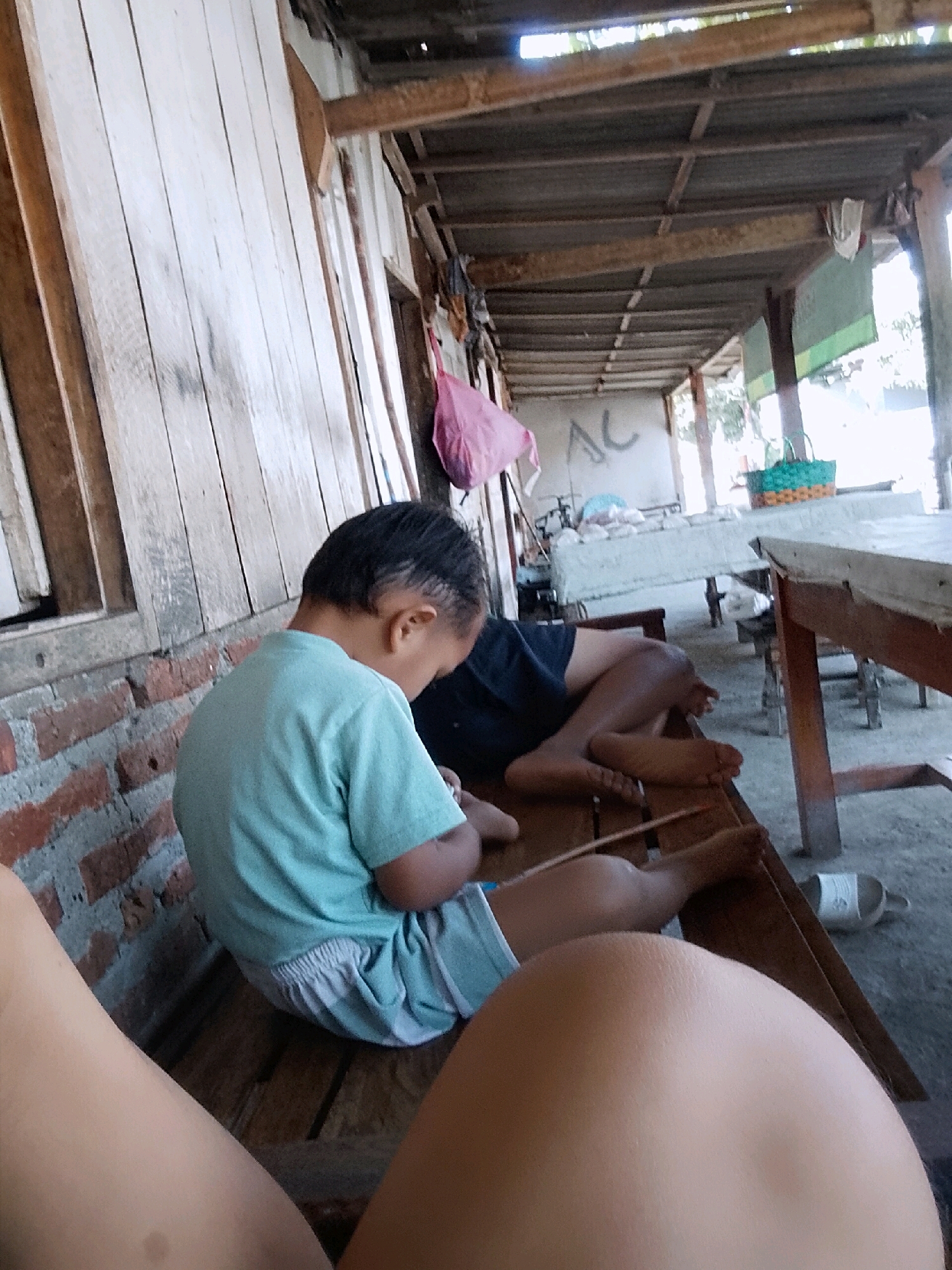 The image depicts a scene in Wadang, Indonesia, likely a rural or semi-rural area, under a covered wooden structure that appears to be an outdoor shelter or extension of a building. The structure has a corrugated roof supported by wooden beams, and the walls are made of vertical wooden planks on the left and brick on the right.
In the foreground, partially visible, are the legs of a person, suggesting the viewpoint of someone sitting or lying down. In the midground, a young child is seated on a wooden bench, wearing a light blue t-shirt and light blue and white striped shorts. The child is looking downwards, and their back is to the camera. To the right of the child, another person, likely an adult, is also seated on a wooden bench, with their legs extended. This person is wearing dark shorts and appears to be barefoot.
The environment is rustic. Behind the people, there are tables covered with what appear to be cloths or tarps. On one table, a green woven basket is visible. Various objects, including what looks like clothing or bags, are hanging from the structure's beams in the background. The ground is dirt or concrete. The overall impression is one of a simple, outdoor living or gathering space.