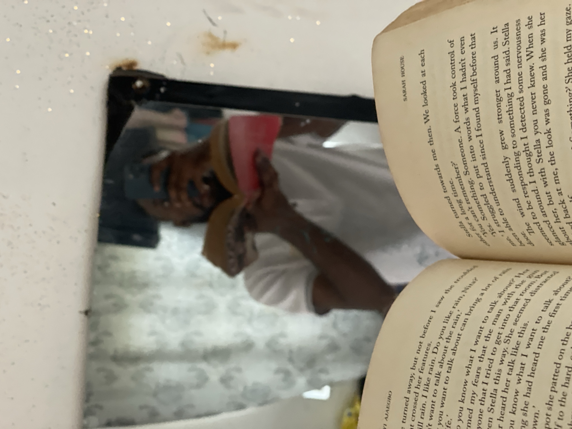 The image shows a reflection in a mirror of a person holding a book. The reflection reveals a dark-skinned individual wearing a light-colored shirt. They are holding the book up to the mirror so the text on the pages is visible. The book's title page shows the name "SARAH HOUSE" and mentions Stella, indicating the book is likely fiction. Another visible name is "YI AJAEGBO". The background is indistinct but suggests a patterned surface, possibly a wall or wallpaper. There is no indication of the location being Lokoja, Nigeria.