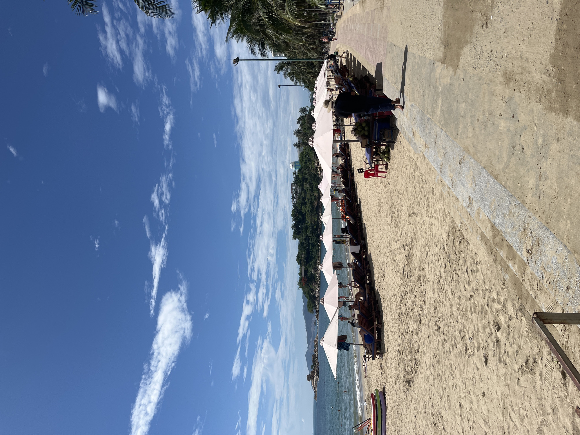 The image captures a vibrant, sunny day at a beach in Bac Nha Trang Ward, Vietnam. The scene is oriented vertically, showing a wide expanse of clear blue sky with scattered white, wispy clouds on the left.
In the foreground and midground, a sandy beach stretches along the coastline, populated by several white beach umbrellas providing shade. Beneath these umbrellas, people are seen relaxing on lounge chairs. Towards the right, a paved promenade or walkway runs parallel to the beach. A street vendor's stall is visible on the sand, adjacent to the walkway, featuring a display of fresh coconuts and red plastic chairs. One person stands near this stall, casting a distinct shadow on the sand and walkway, suggesting a bright sun high in the sky.
Beyond the beach umbrellas, the calm, blue-green ocean stretches out, with hints of distant land and hills covered in lush greenery. Numerous palm trees line the promenade, adding to the tropical ambiance. The overall atmosphere is one of leisure and relaxation, indicative of a typical beach holiday destination.
The time of day appears to be late morning or early afternoon, given the strong sunlight and shadows. The weather is clear and pleasant. There is no discernible text visible in the image.