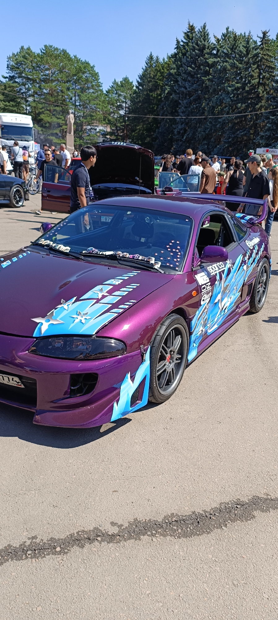 On a clear, sunny day, a bright purple sports car with blue and white graphics is parked on an asphalt surface. The car has a sleek design with a spoiler and distinctive headlights. The graphics on the car include abstract shapes and what appear to be stars and flames. The car is adorned with various stickers, including "Sparco" and "OZ Racing." In the background, a crowd of people are gathered, some looking at the car and others milling about. Trees and a blue sky form the backdrop. There is also a white truck visible in the distance. The ground has a visible crack running through it. The scene appears to be an outdoor car show or exhibition.
