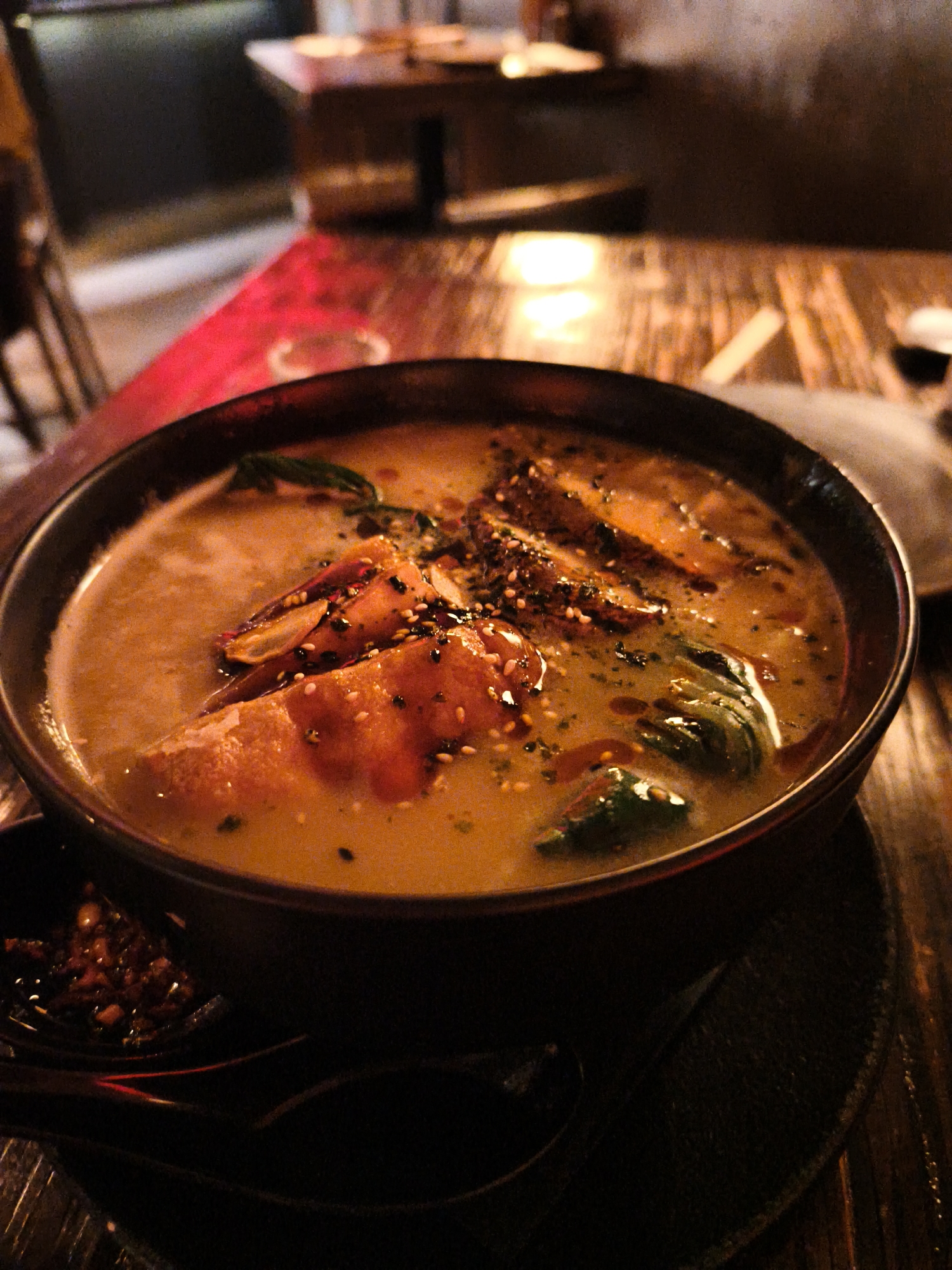 A close-up, eye-level shot features a dark, rustic bowl of ramen soup situated on a dark wooden table. The ramen is a creamy, light orange color, with visible pieces of protein and leafy greens. Grains of sesame seeds and other seasonings are sprinkled across the surface of the dish. To the lower left of the bowl, a small dish containing a chili-based condiment is visible, along with two dark spoons resting on the table. The background is blurred and dark, suggesting an indoor setting with low lighting. The ambient light creates a warm, inviting atmosphere. The scene is located in Be'er Sheva, Israel, likely within a restaurant environment.