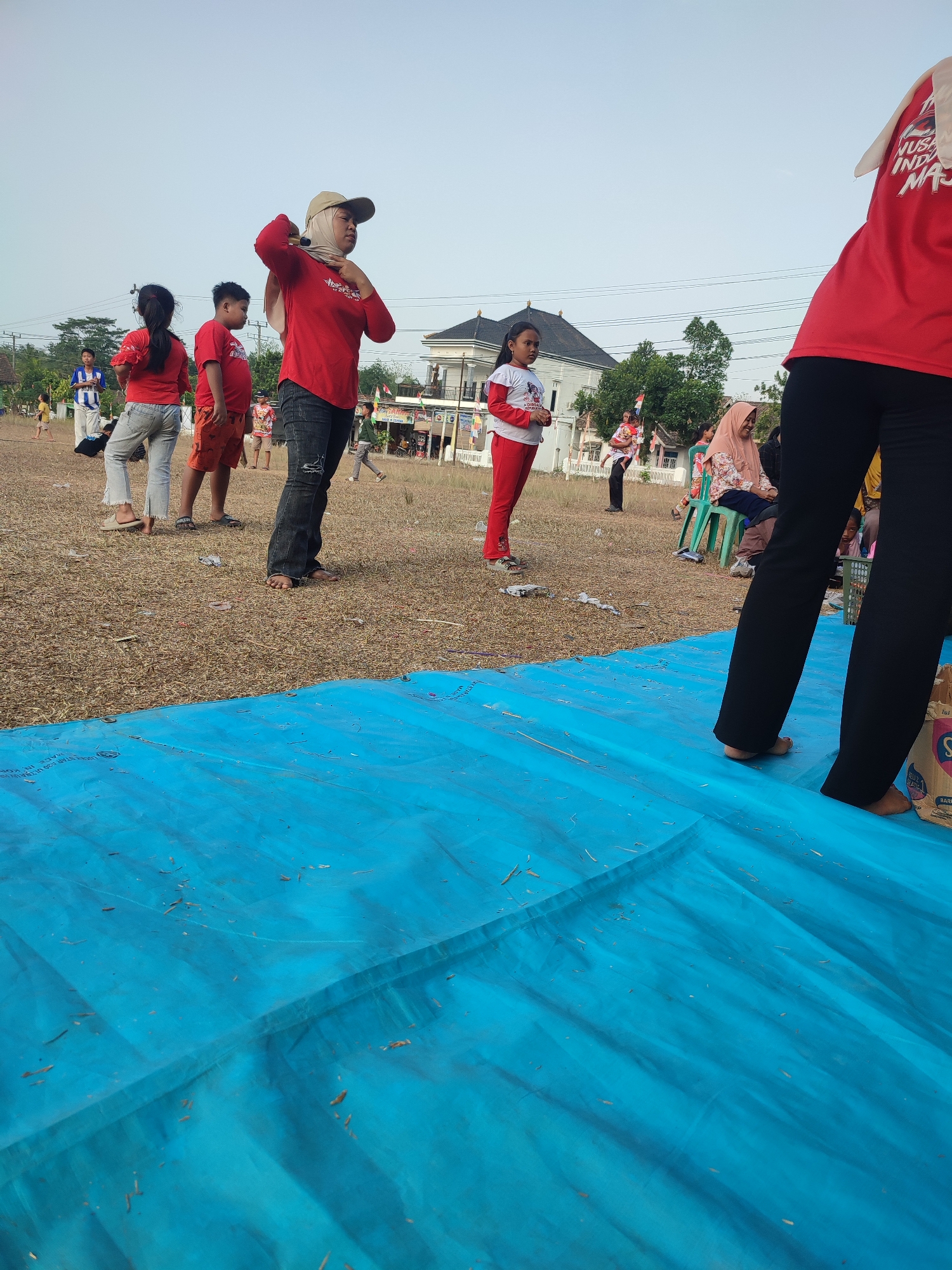 A group of people, including children and adults, are gathered outdoors in what appears to be a dry, grassy field with some debris scattered on the ground. The scene takes place in front of a residential area, with houses visible in the background. The sky is clear and pale, suggesting daytime. A large blue tarp covers the foreground.
In the foreground, to the right, the lower half of a person wearing black pants is visible. To the left, a large blue tarp is spread out, with some dry grass and leaves on its surface.
In the middle ground, several individuals are positioned. A woman wearing a beige headscarf and a red shirt is standing, with her right hand raised towards her head. Next to her, to her left, are two children, a boy and a girl, also wearing red shirts. The boy is wearing orange shorts, and the girl is wearing light blue jeans. Further to the left, another child in a red shirt is visible.
In the background, more people are dispersed. A person in a white and blue shirt is standing, and other individuals are seen walking or standing. Behind the people, there are houses with distinctive architecture, including what appears to be a white, multi-story building with a dark roof. Trees and utility poles are also present. The overall impression is of a community gathering or event taking place in an open, outdoor space.