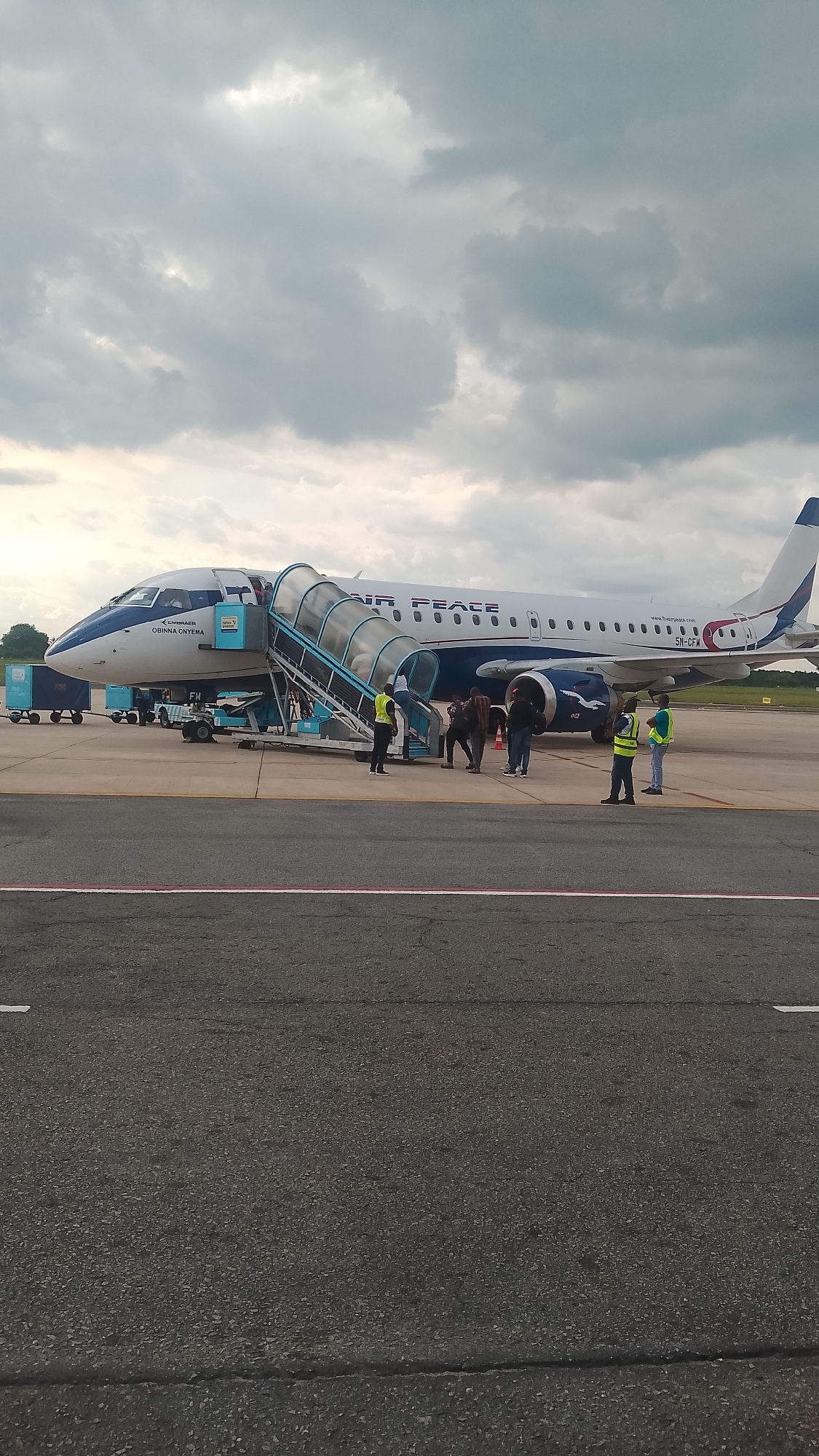 The image shows an Air Peace Embraer ERJ-145 aircraft parked on an airport tarmac. A mobile stairway is attached to the aircraft's forward exit, and several people appear to be boarding or deplaning. Two ground crew members wearing yellow safety vests stand near the wing of the aircraft. Other ground support equipment is visible near the front of the plane. The sky is overcast. There is no indication the location is Orerokpe, Nigeria.