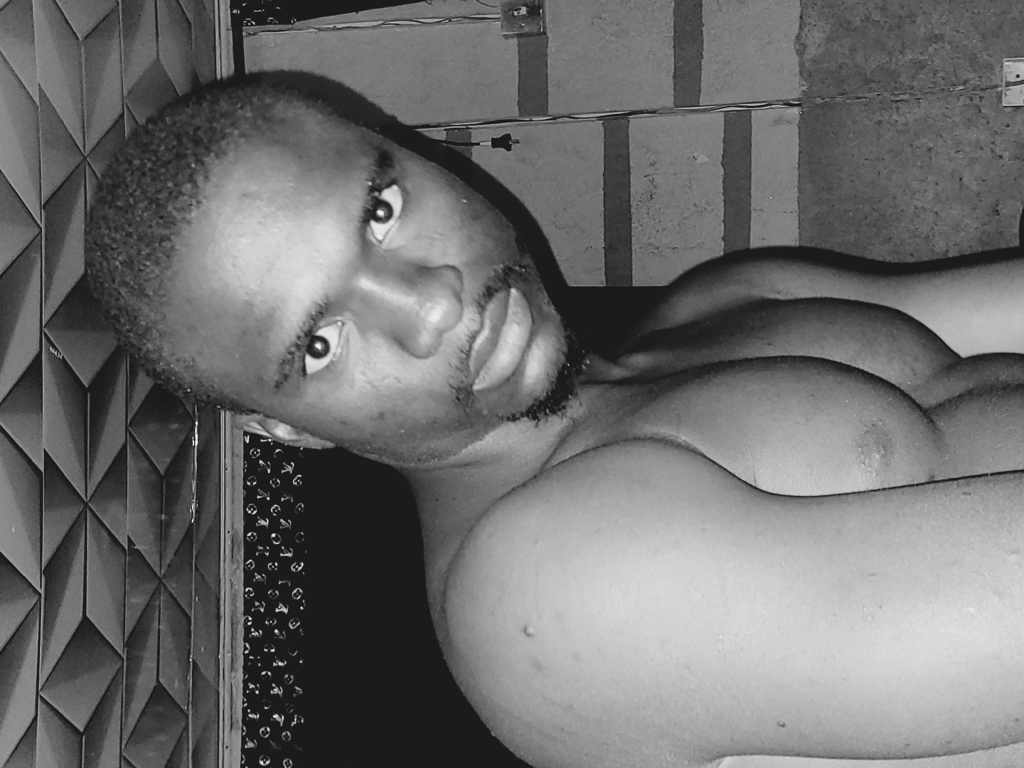 A monochrome, medium shot depicts a muscular, shirtless black man with a close-cropped haircut and a short beard. He is looking directly at the camera with a serious expression. The background is a mix of patterned walls and a concrete structure with visible electrical wiring and outlets. There's no indication in the image that places it specifically in Bukuru, Nigeria.