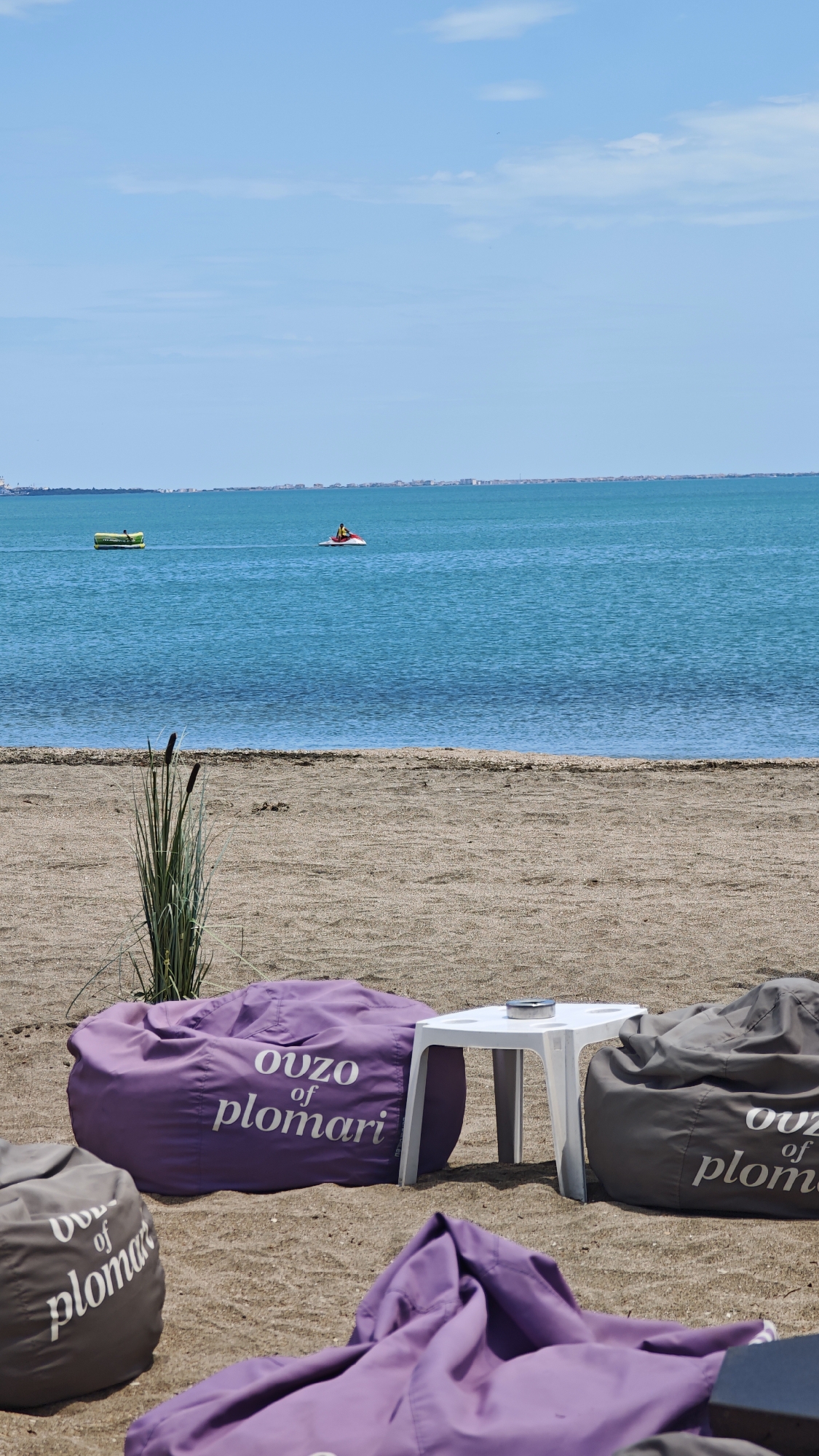The image shows a beach scene. In the foreground, there are beanbag chairs with the words "ouzo of plomari" on them and a small white plastic table. In the middle ground, there's a sandy beach with some vegetation. In the background, there is the water of the Black Sea. A yellow inflatable and a jet ski are floating on the water. On the horizon, there's a cityscape which is of Burgas, Bulgaria. The sky is blue with a few clouds.