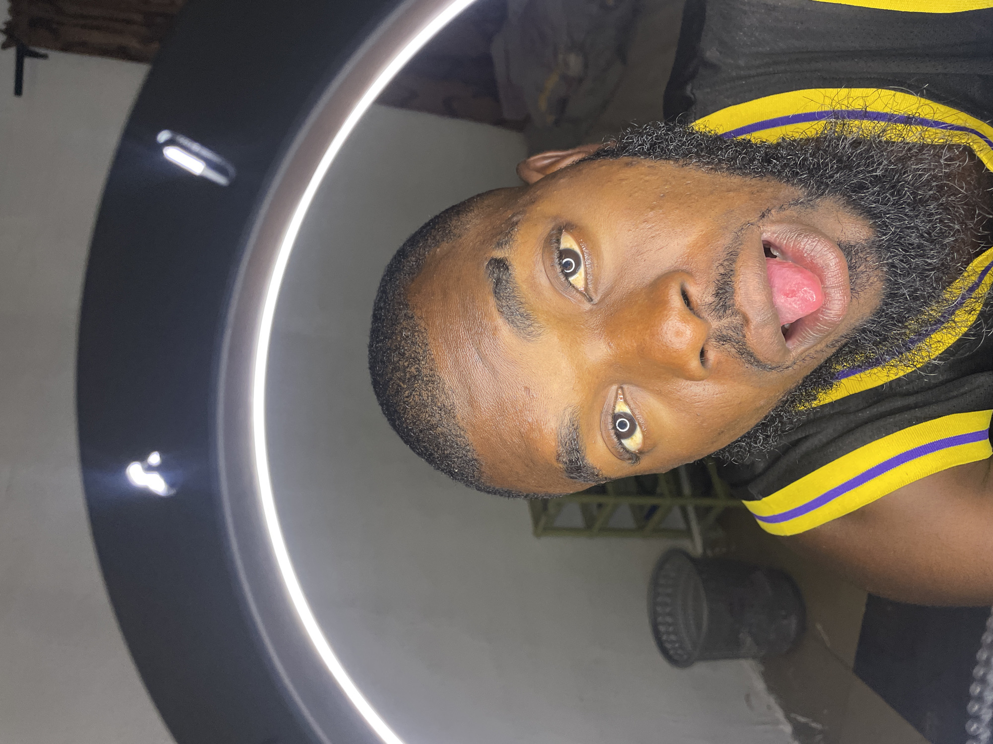 A close-up selfie of a Black man with a full beard and a dark complexion is captured from the chest up. The man is wearing a black jersey with yellow and purple stripes on the collar. His mouth is open with his pink tongue extended, and his eyes are looking directly at the camera. A ring light is visible in the background, creating a bright circular reflection in his right eye. To the left of the man, there is a dark gray wastebasket. Further in the background, a yellow shelf unit is partially visible. The scene appears to be indoors, possibly in a residential setting in Jos, Nigeria.